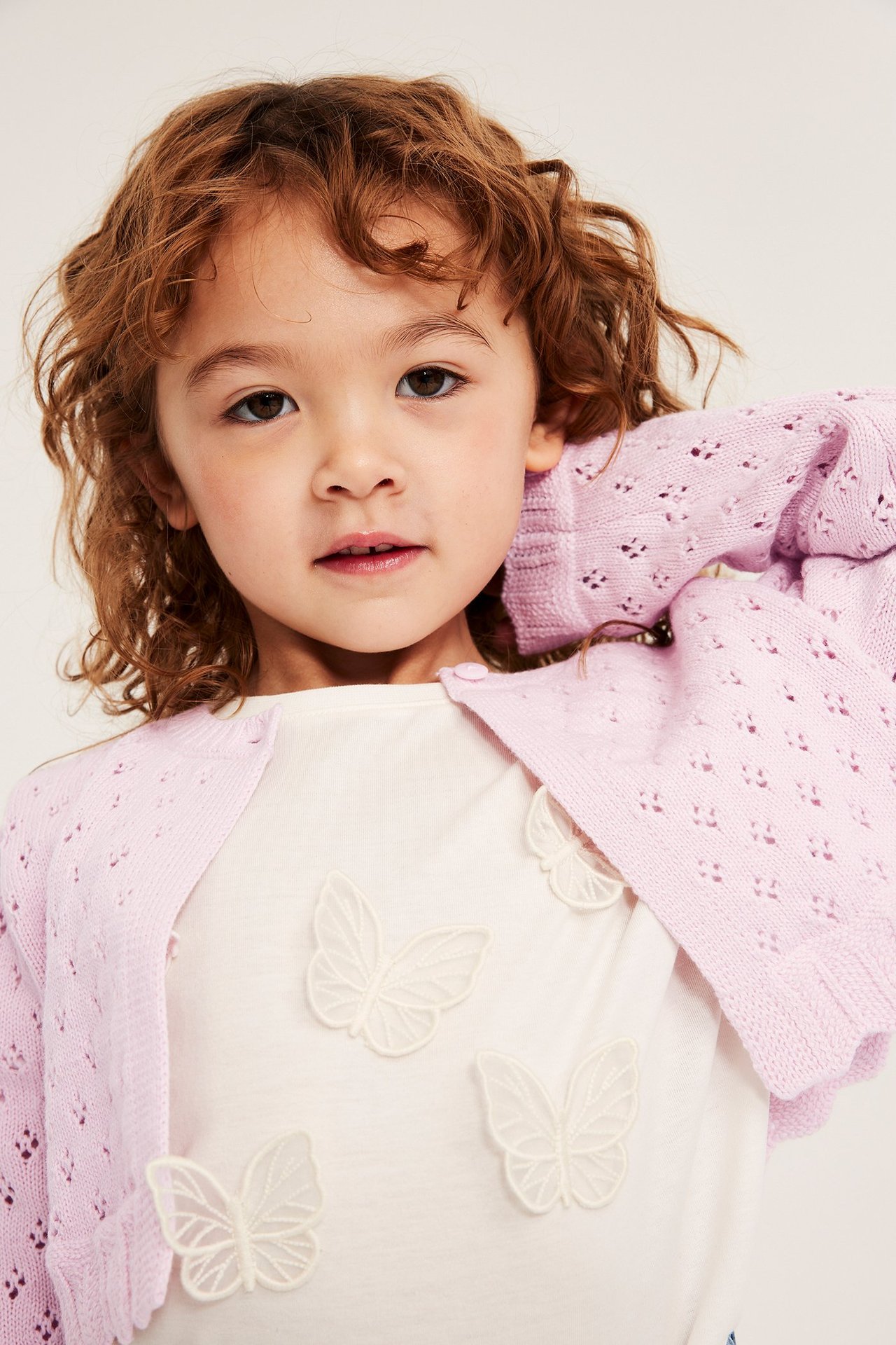 Närbild framifrån på ett barn i en ljusrosa mönsterstickad bomullskofta över en vit t-shirt med fjärilsapplikationer.