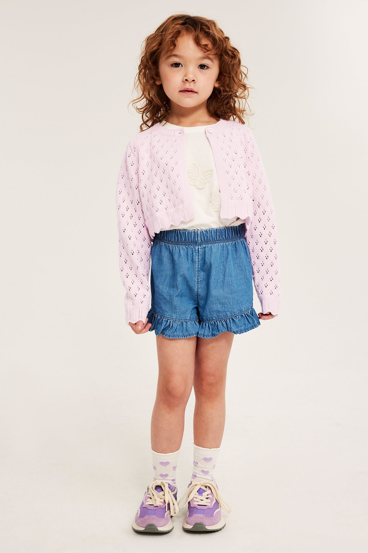 Edestä: Lapsi yllään vaaleanpunainen puuvillaneuletakki, valkoinen t-paita, siniset shortsit, sydänsukat ja violetit lenkkarit.