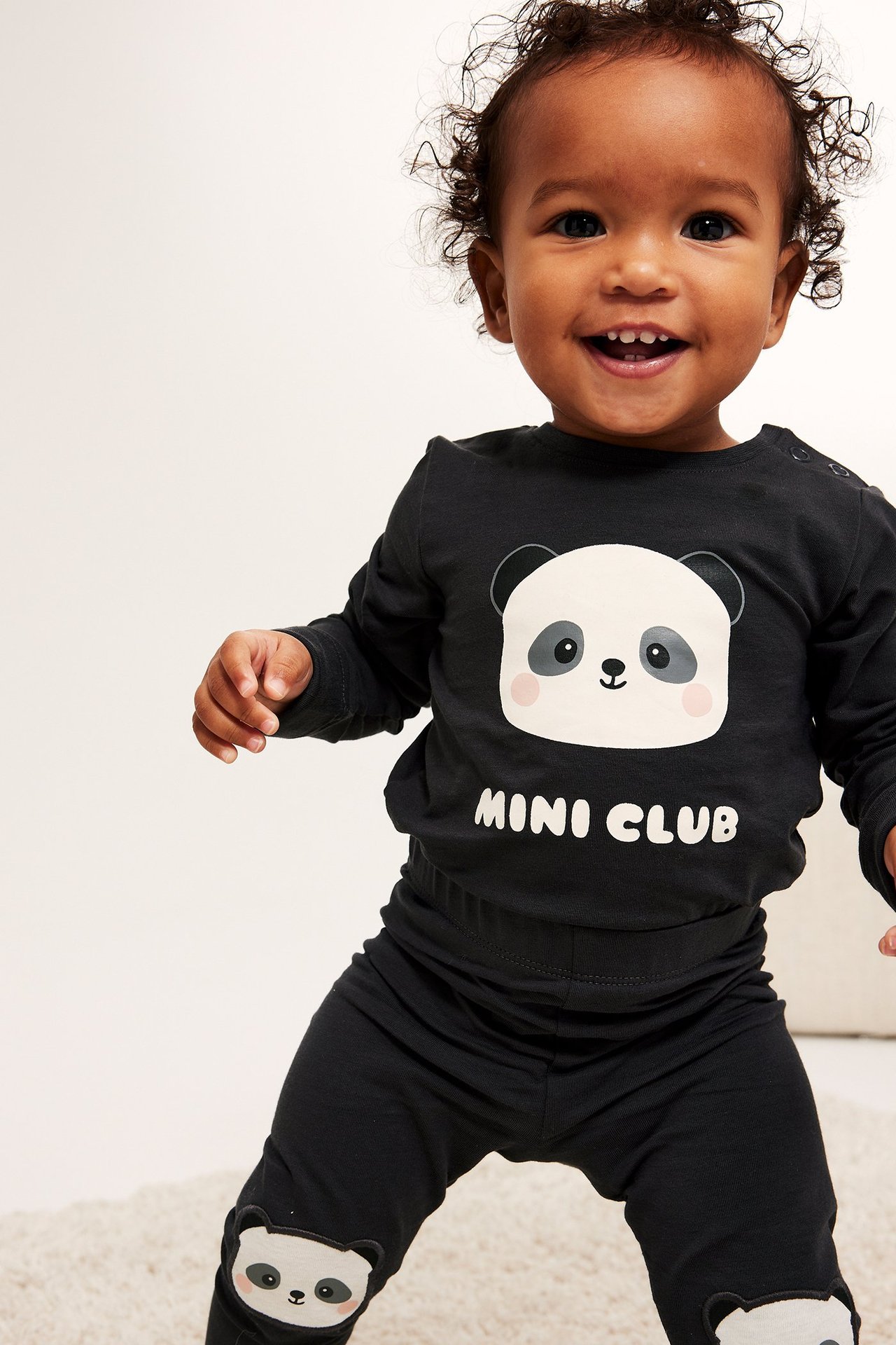 Smilende baby, forfra, i svart langermet pandamønstret body og svarte leggings med pandapatches.