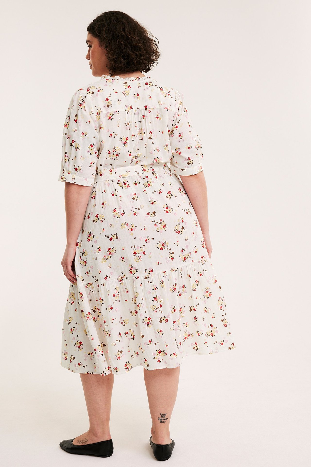 Bakifrån på kvinna i offwhite mönstrad bomullsmidiklänning med puffärmar, knytskärp och svarta platta skor.