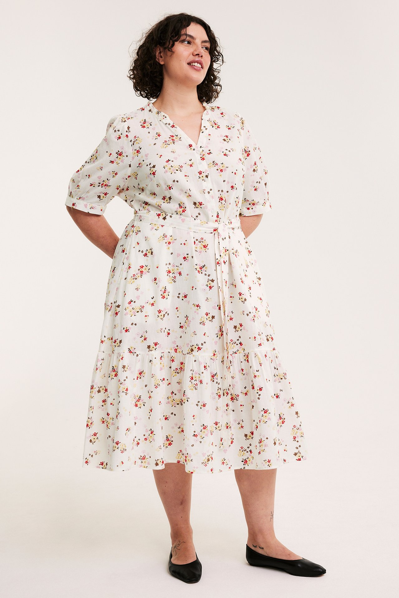 Front view: kvinna i offwhite mönstrad bomulls-midiklänning med puffärmar, knytskärp och svarta ballerinaskor.