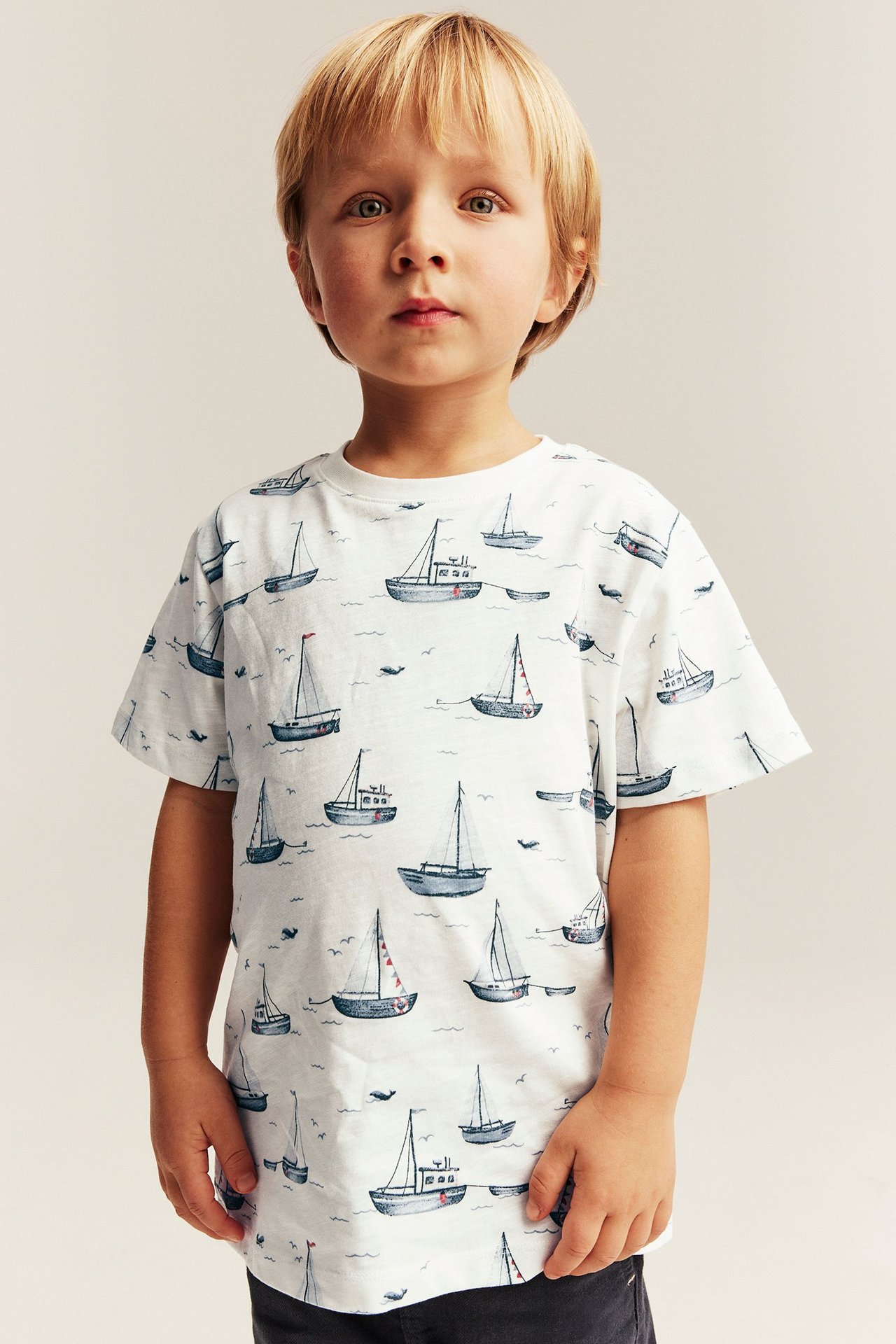 Framifrån, ett barn i en offwhite kortärmad bomulls-t-shirt med segelbåtsmönster.