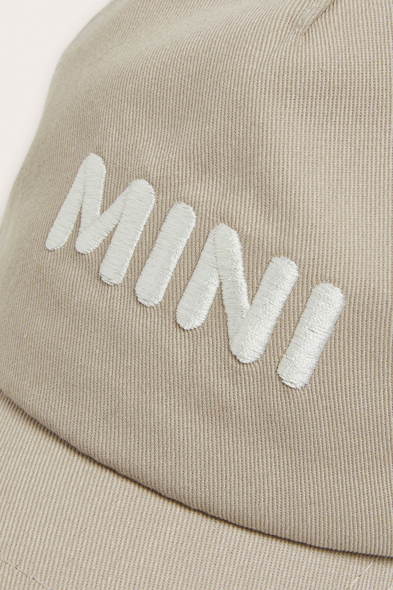 Nærbilde av en beige bomullscaps med hvit «MINI»-broderi foran.