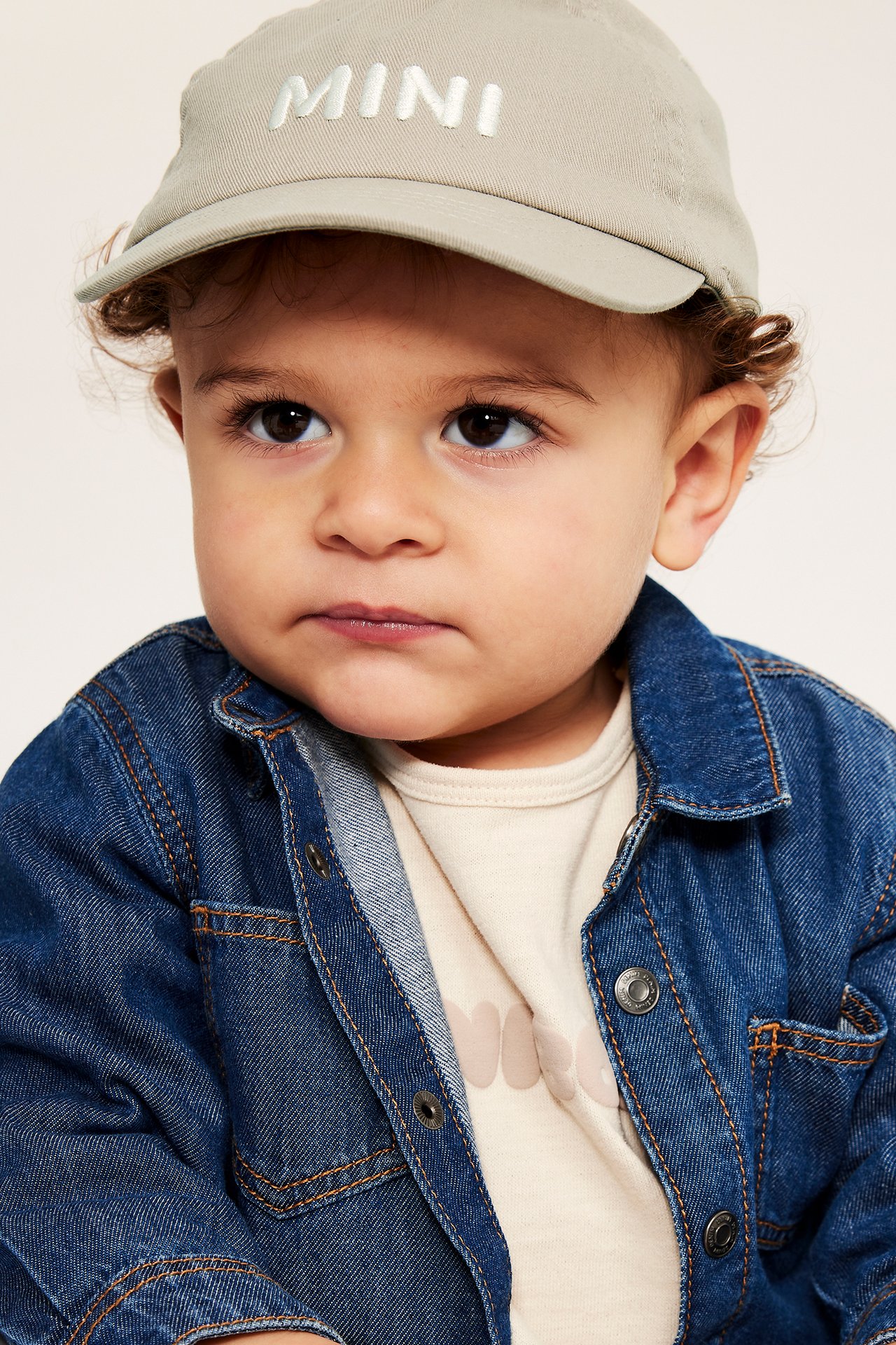 Nærbilde av en baby med beige økologisk bomullscaps med broderi og en blå dongerijakke.