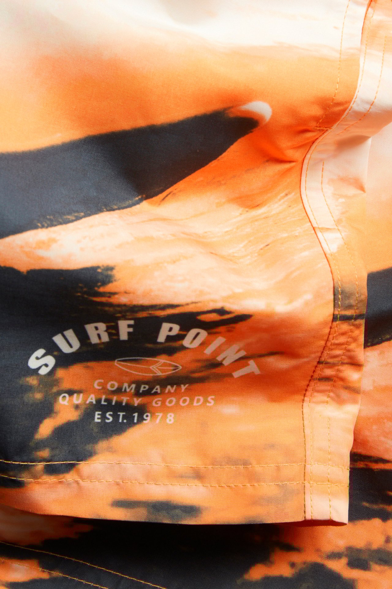 Närbild av orange vävda badshorts med mörkt surfprint och vit 'SURF POINT'-logga.
