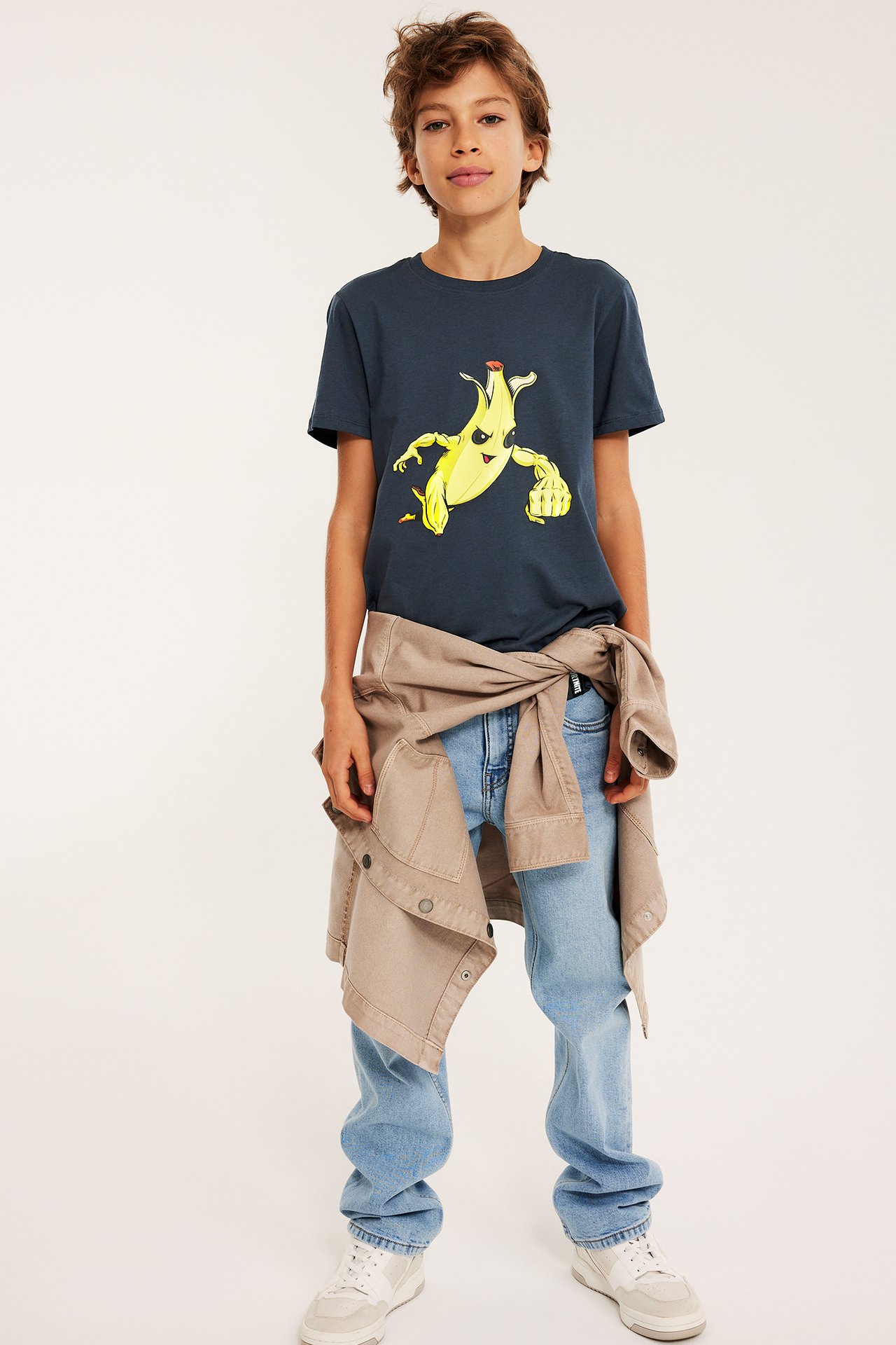 Barn bär blå Fortnite T-shirt med banantryck, ljusblå jeans, beige jacka, vita sneakers.