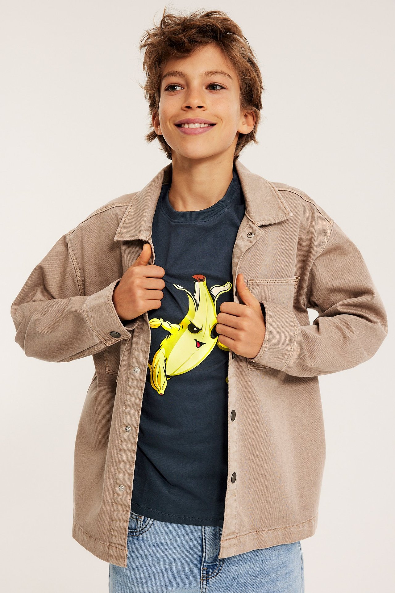 Widok z przodu dziecka w niebieskiej koszulce Fortnite z bananem, pod otwartą beżową koszulą i dżinsami.