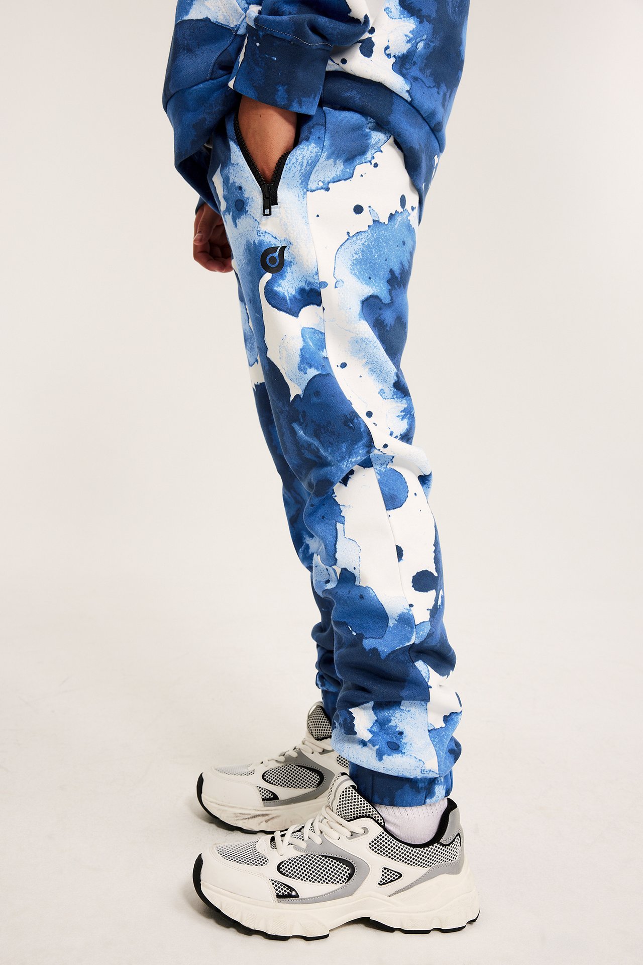 Widok z boku dziecka w biało-niebieskich spodniach dresowych z kieszenią na zamek, białych skarpetach i trampkach.