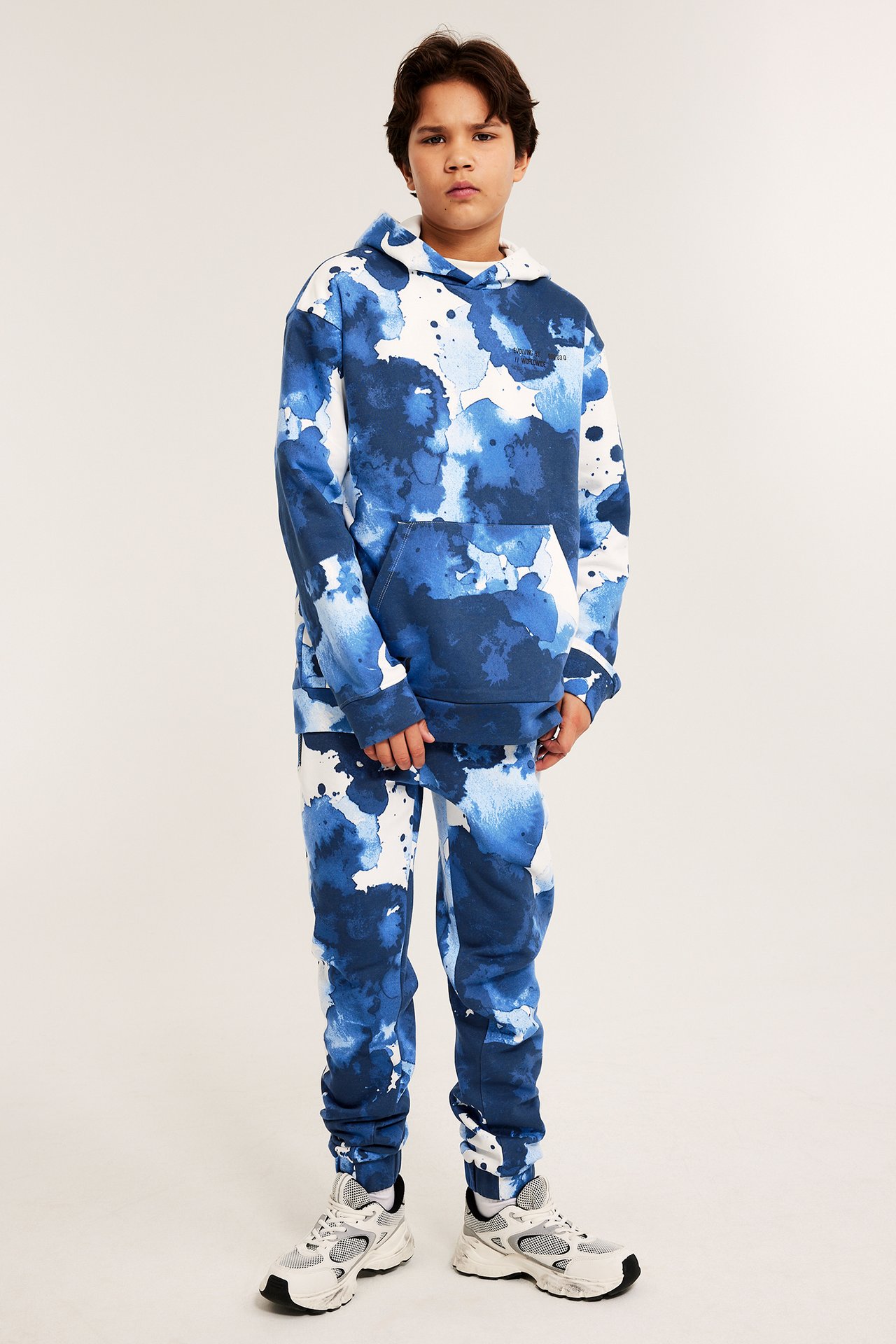 Widok z przodu dziecka w off-white/niebieskich wzorzystych dresach, pasującej bluzie i trampkach.
