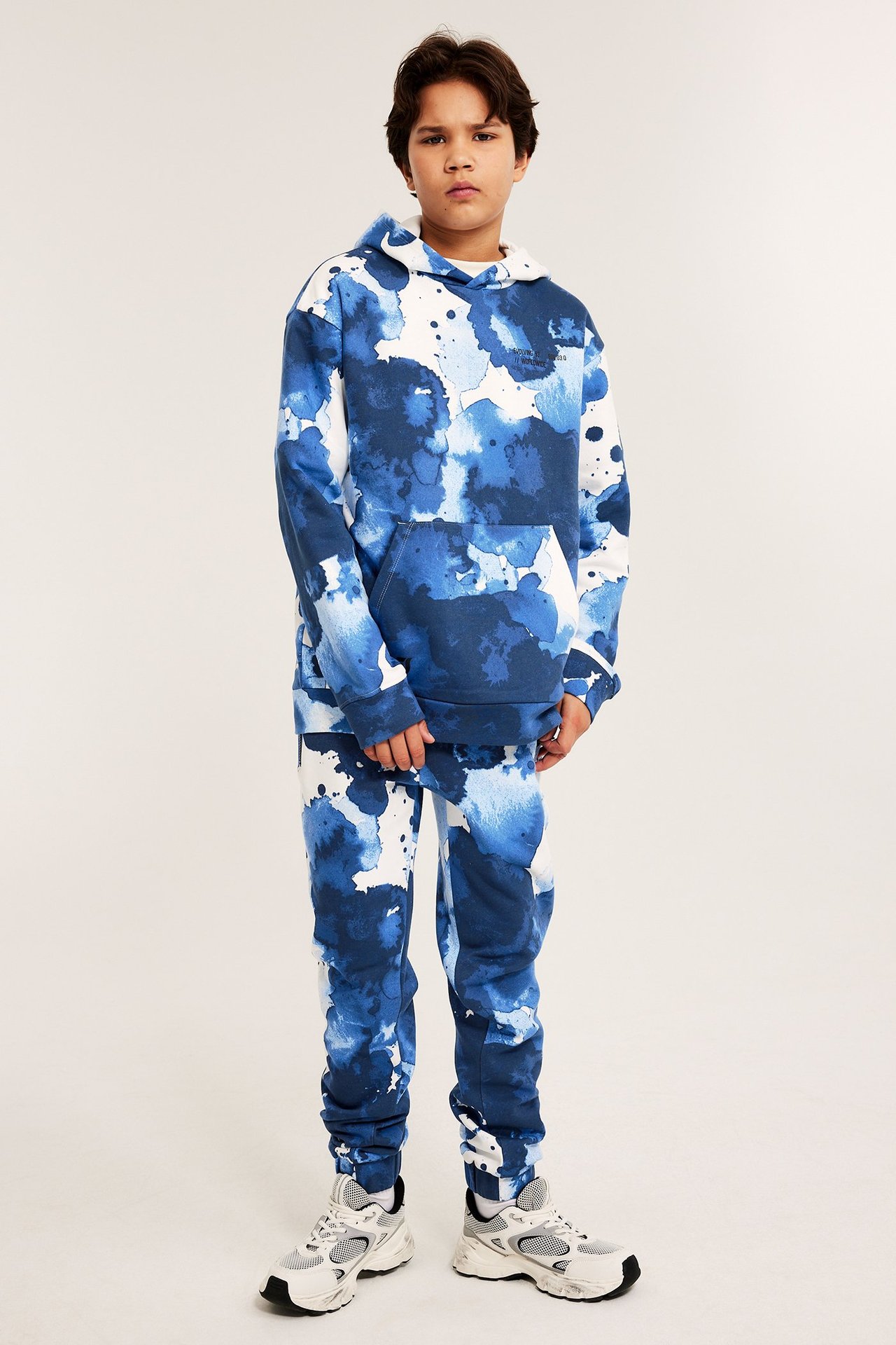 Widok z przodu dziecka w off-white/niebieskich wzorzystych dresach, pasującej bluzie i trampkach.