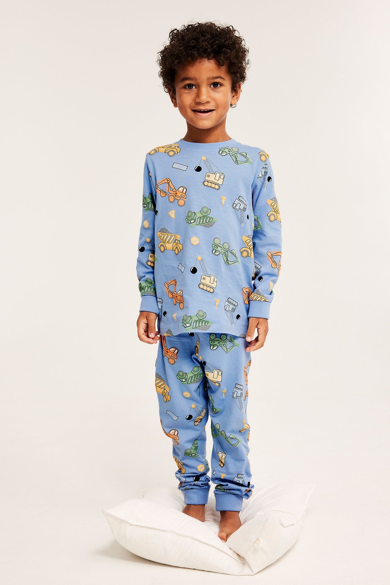 Forfra: Barn i lyseblå bomullspysjamas med anleggskjøretøymønster, står på en hvit pute.