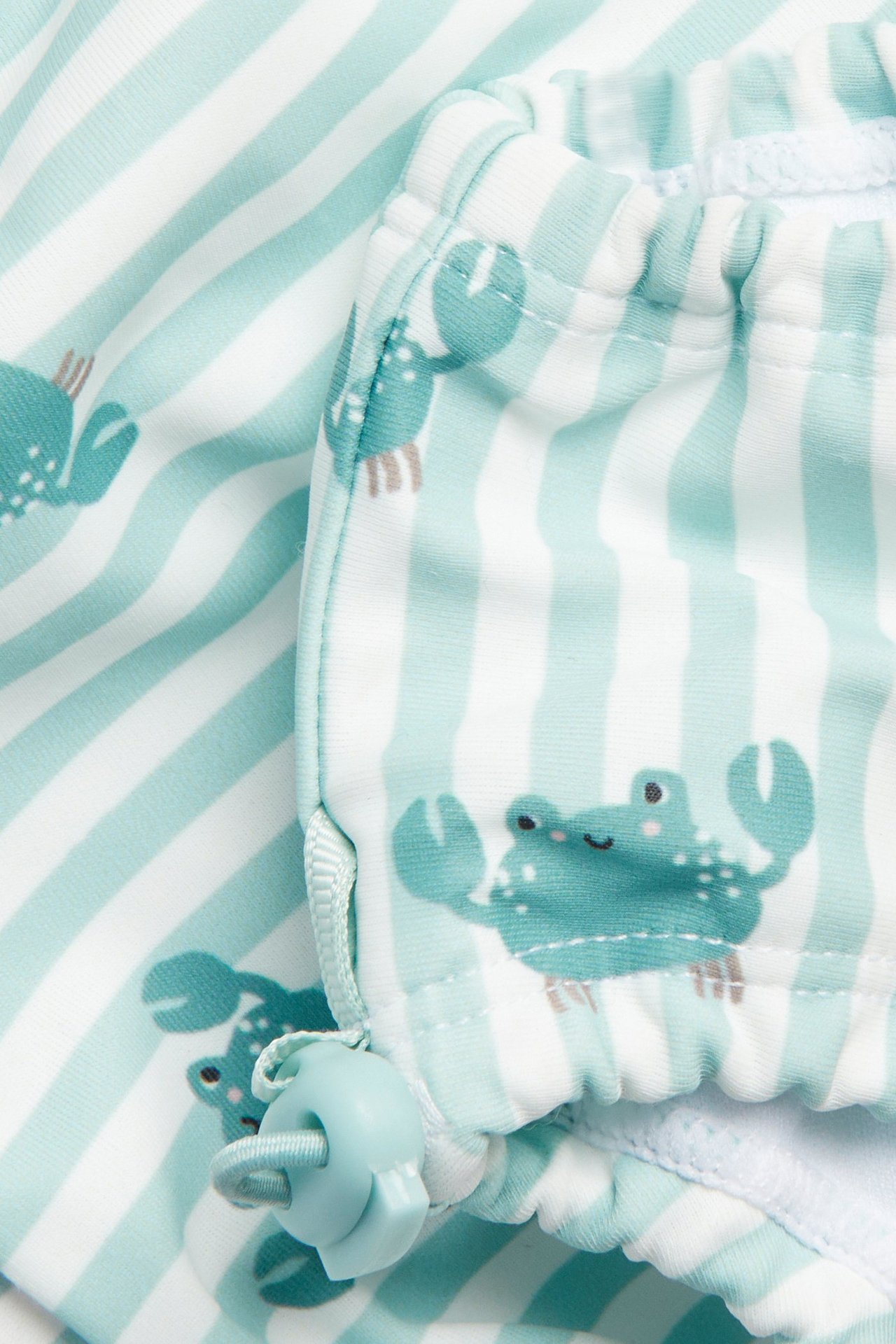 Närbild av babybadbyxor, ljus turkos och vitrandiga, med krabbmönster och justerbar benslutsstopper.