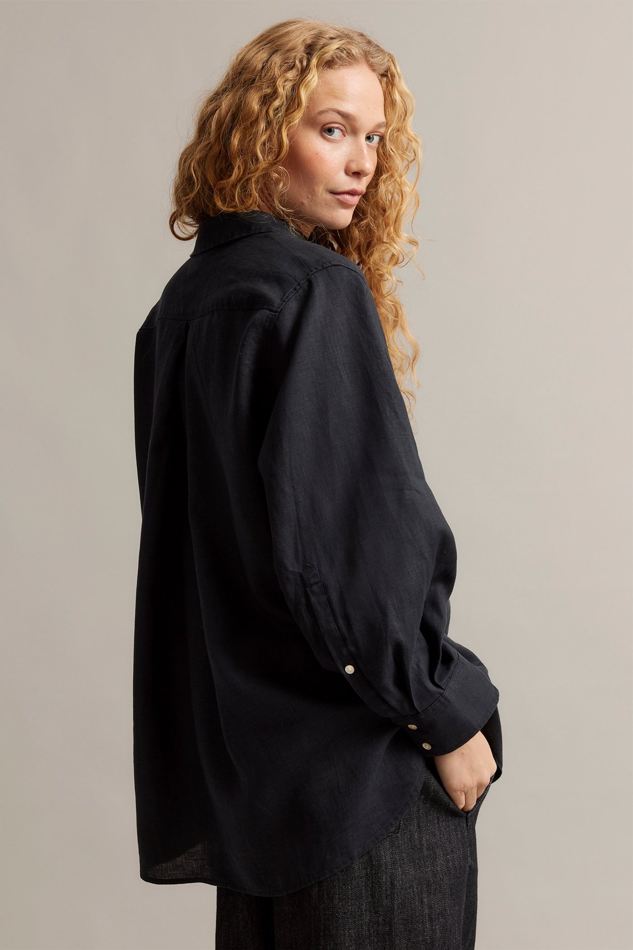 Widok z boku kobiety w czarnej lnianej koszuli oversize z perłowymi guzikami i ciemnymi spodniami.