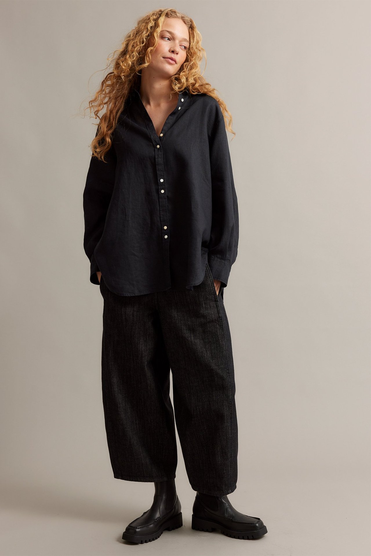 Z przodu: kobieta w czarnej, obszernej lnianej koszuli z guzikami z masy perłowej, ciemnych szerokich spodniach i czarnych masywnych botkach.