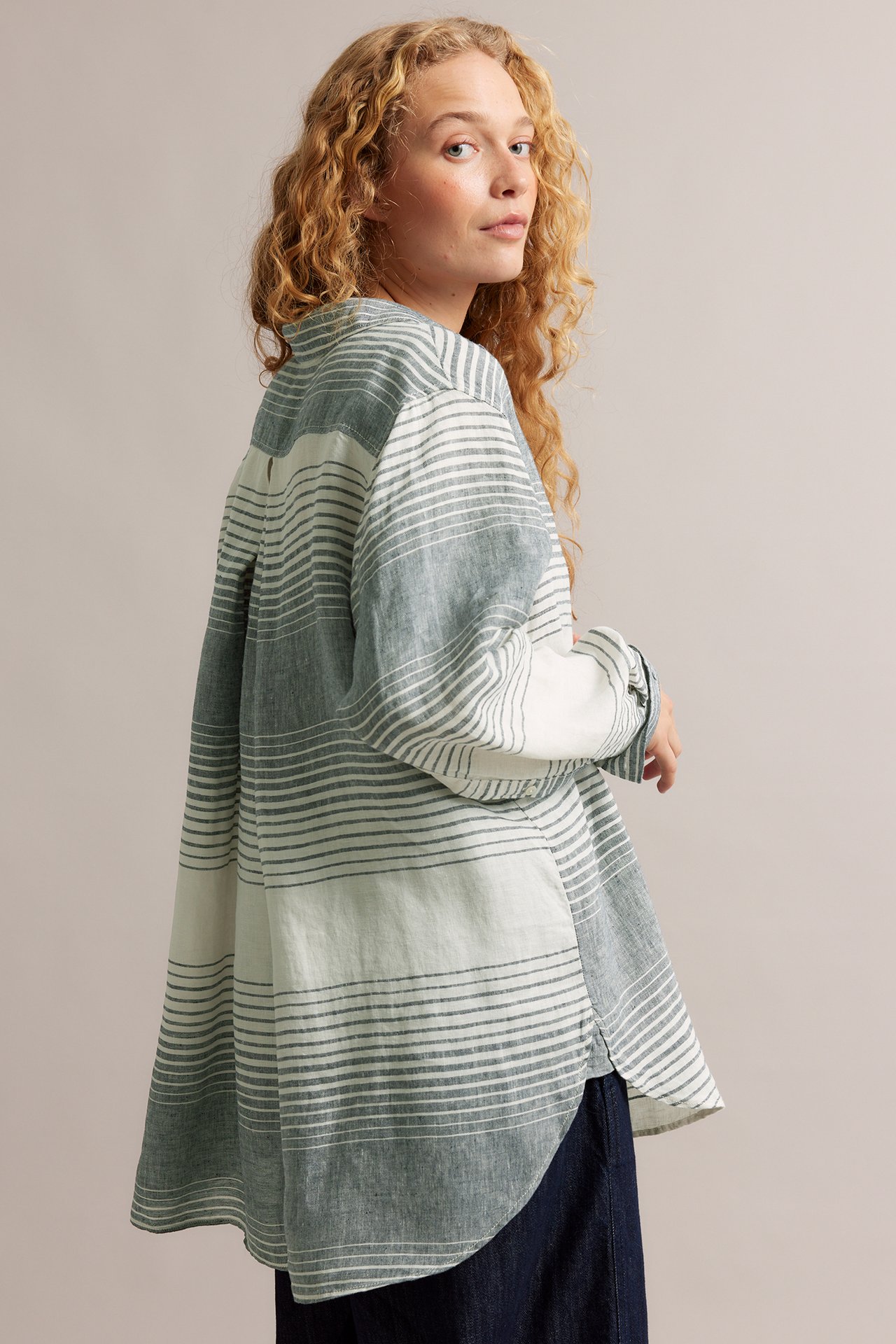 Bakifrån på kvinna i oversizad, blågrå och vitrandig linneskjorta över mörka byxor.