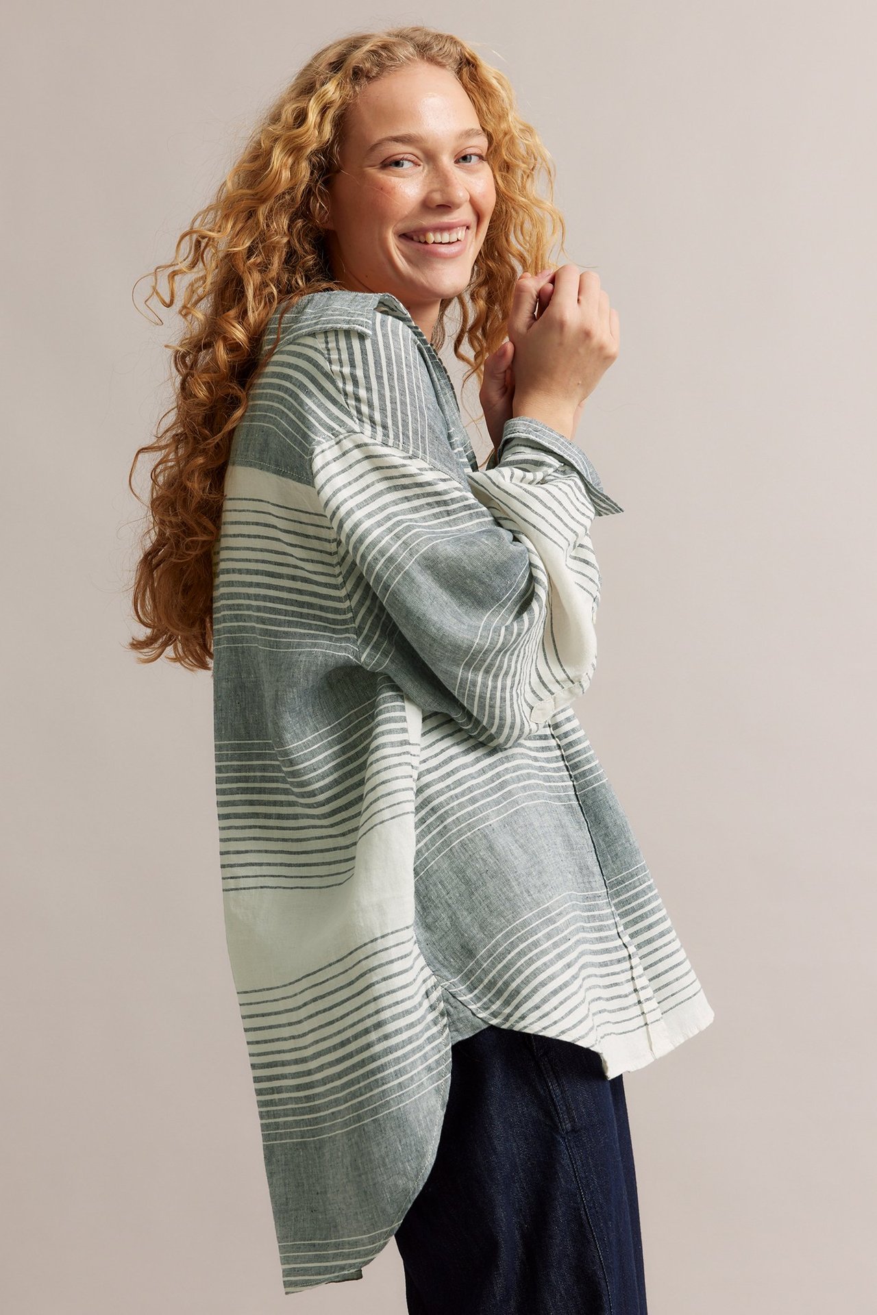 Sidovy av leende kvinna i en oversized mörkblå-vit randig linneskjorta över mörka byxor.