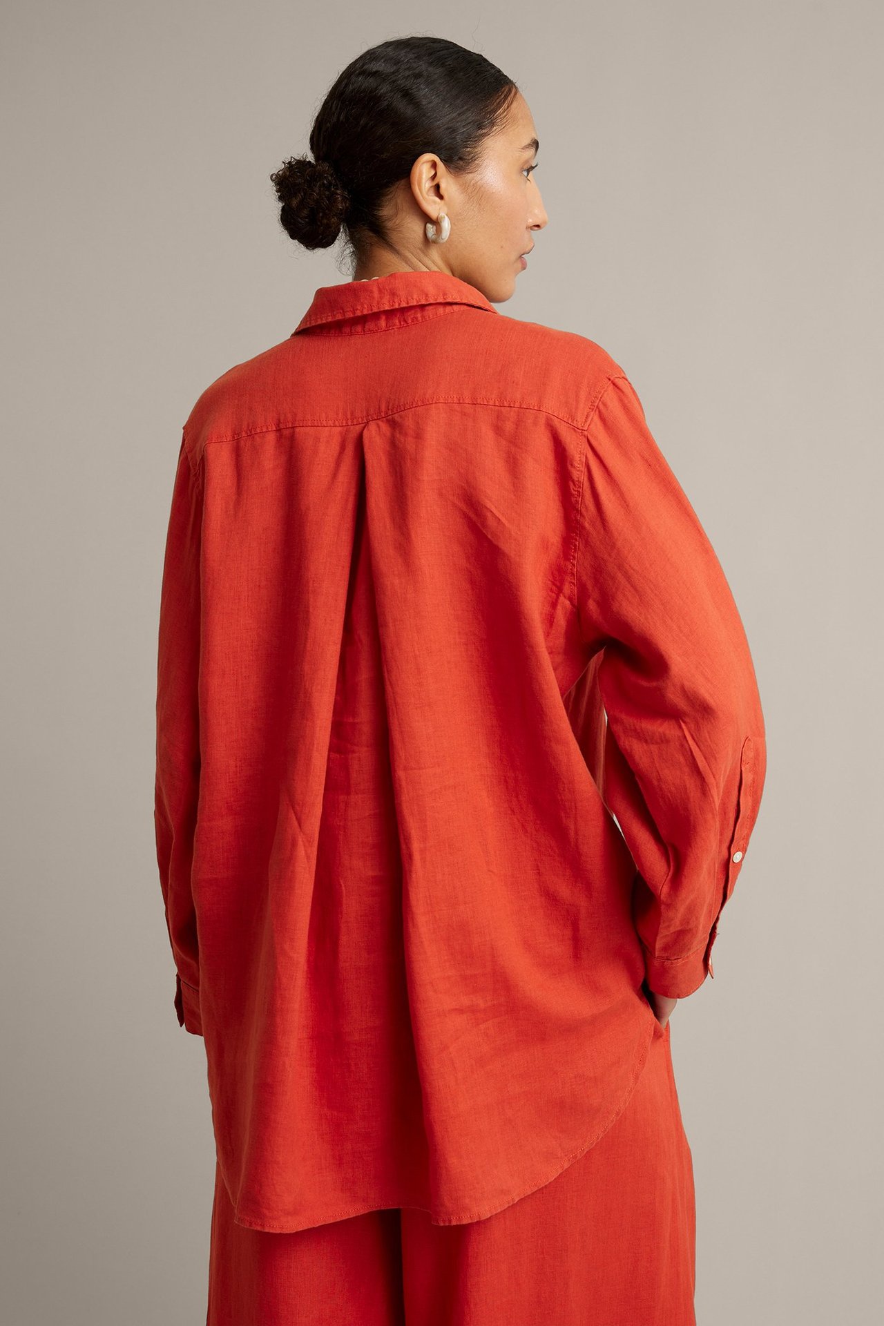 Bakfra: kvinne i oversized rød linskjorte med ryggfold, matchende bukser og øreringer.