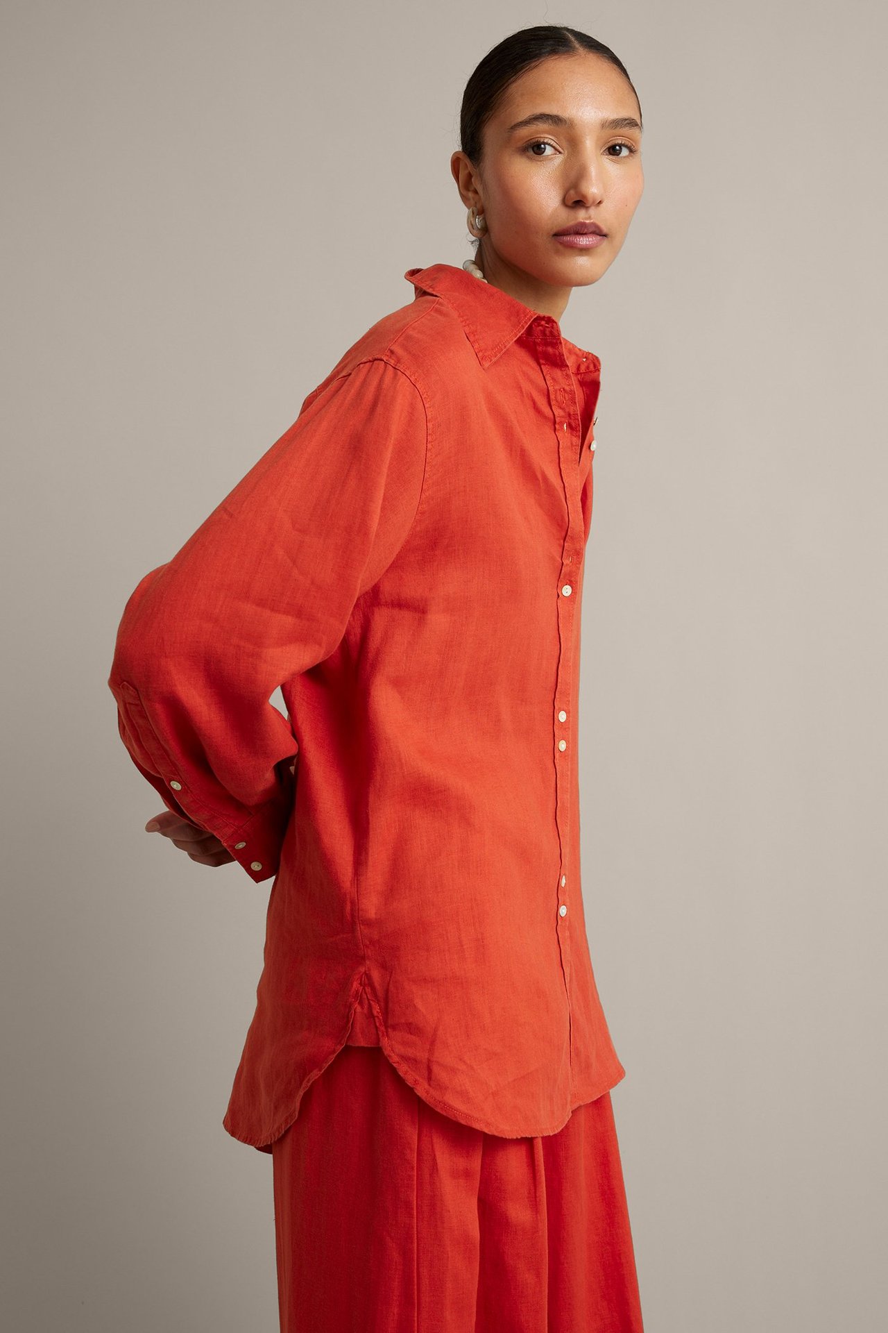 Sidebilde av en kvinne i en rød oversized linskjorte med perlemorsknapper og matchende røde bukser.