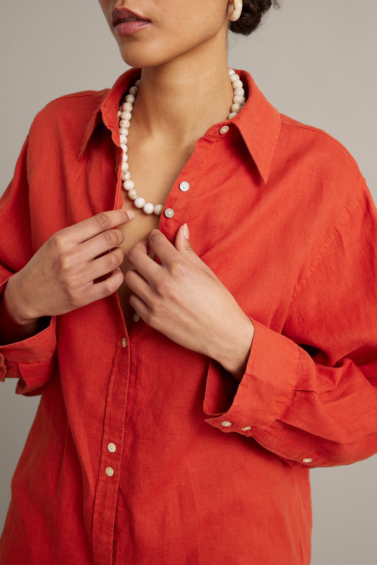 Nærbilde av kvinne i løs, rød linskjorte med perlemorknapper og hvitt perlekjede.