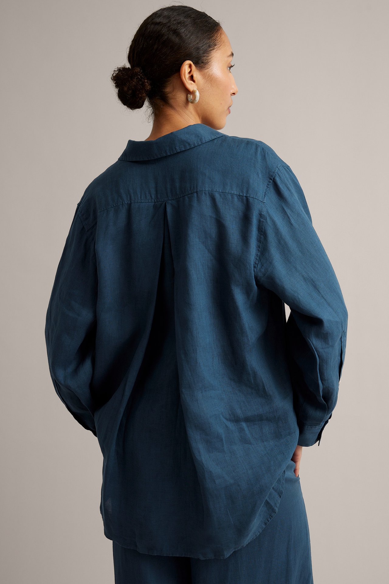 Bakfra, en kvinne i løs, blå linskjorte med ryggfold og matchende bukser.