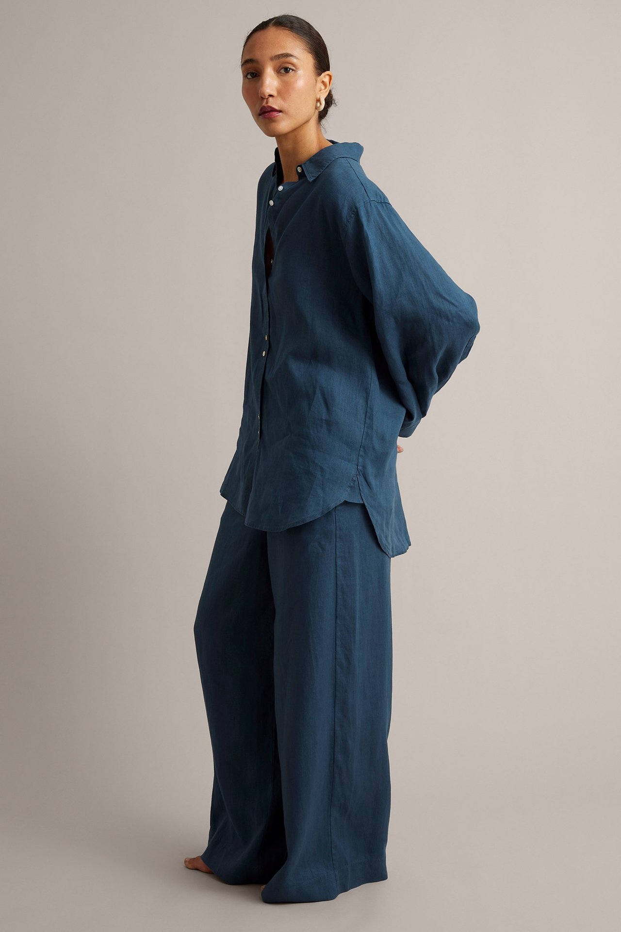 Nainen sivukuvassa oversized sinisessä pellavapaidassa, jossa helmiäisnapit, ja yhteensopivissa leveälahkeisissa housuissa.