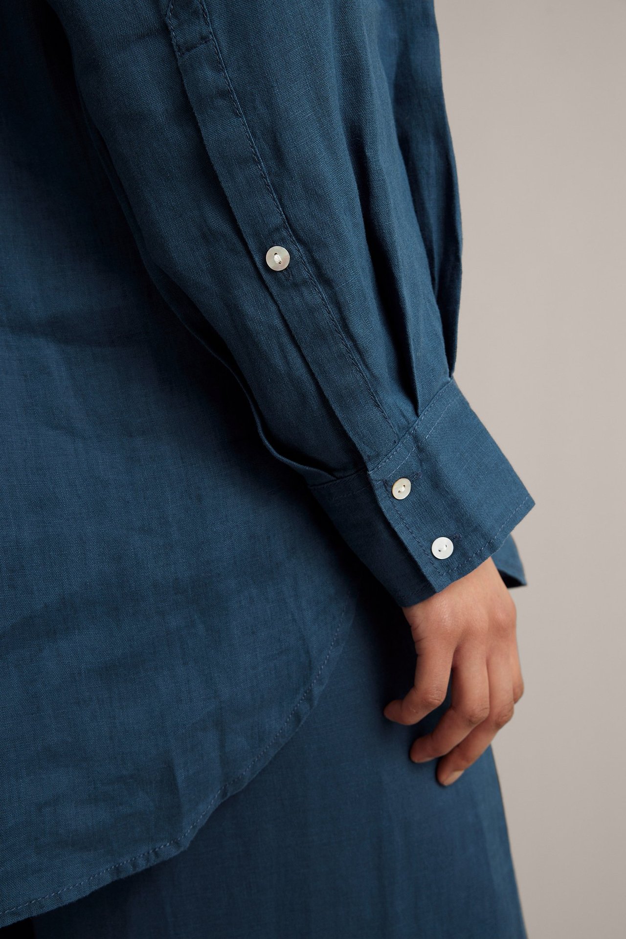 Nærbilde av en kvinnes arm i en blå, oversized linskjorte med hvite perlemorknapper på mansjetten.