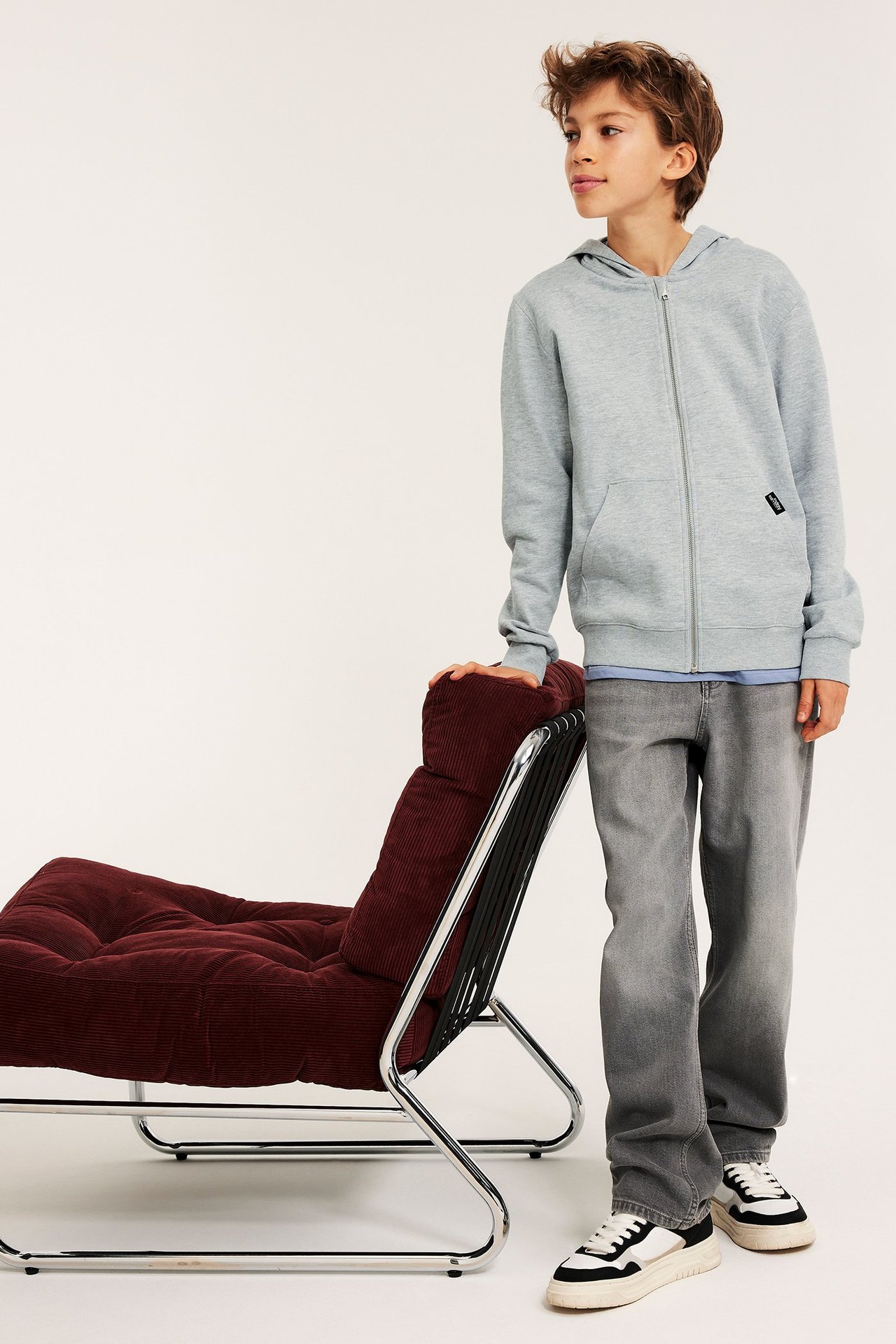 Frontvy: kille i melerad ljusgrå hoodie med dragkedja, blå topp, grå jeans och vita sneakers.