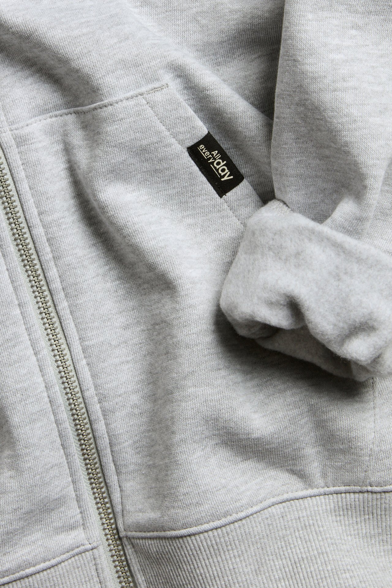 Närbild av en barnhoodie i ljusgråmelerad bomull med dragkedja, känguruficka och ribbad mudd.