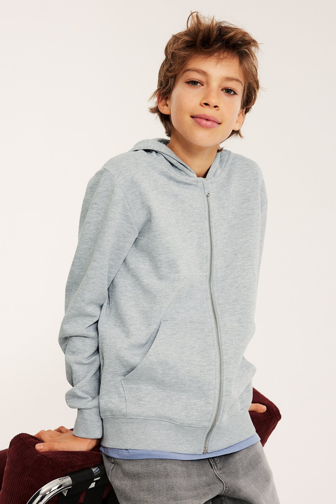 Framsida: Barn i ljusgrå melerad hoodie med dragkedja, kängurufickor, blå topp och grå jeans.