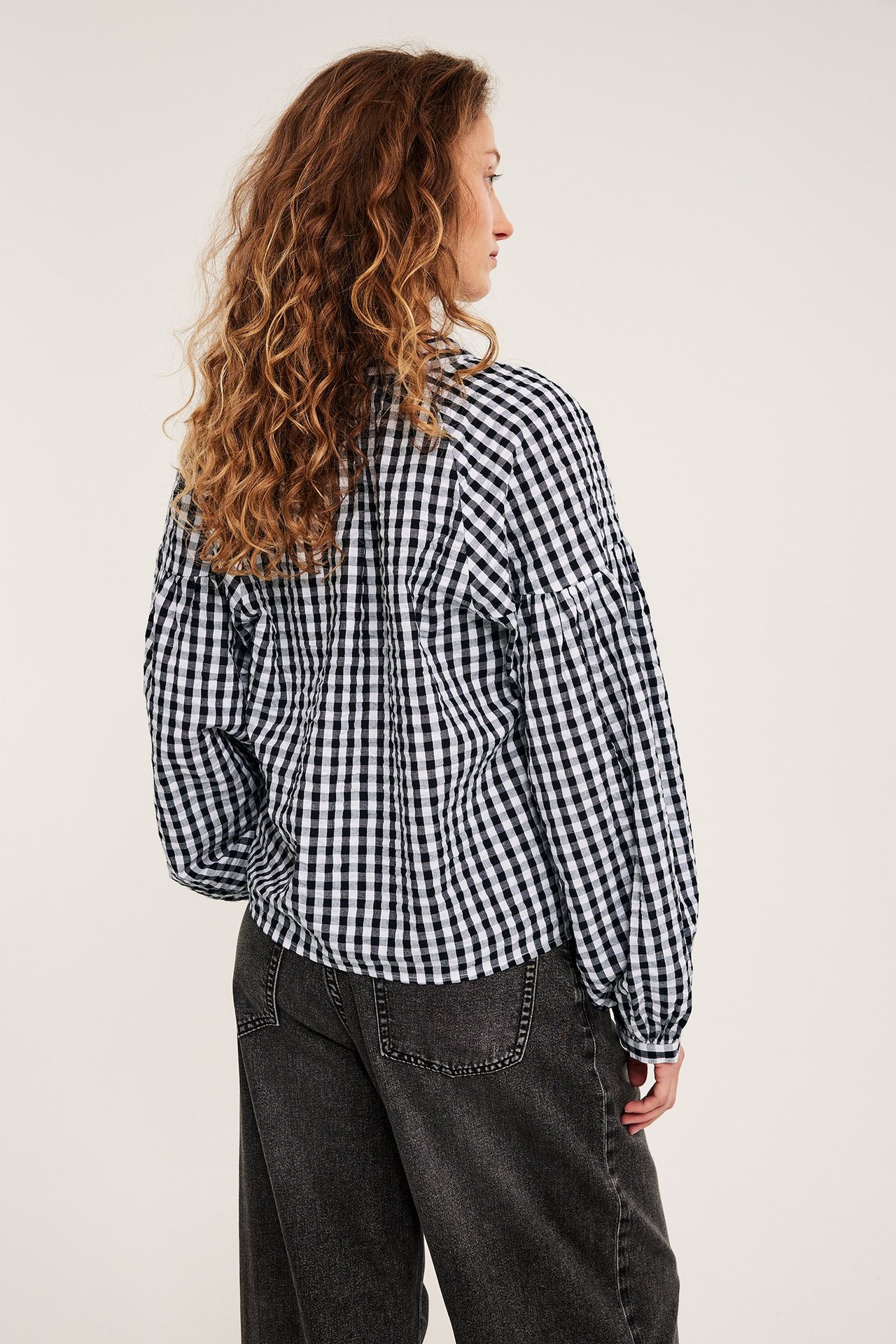 Kvinne i svart-hvitrutet seersuckerbluse med lange ermer og mørke jeans, bakfra.