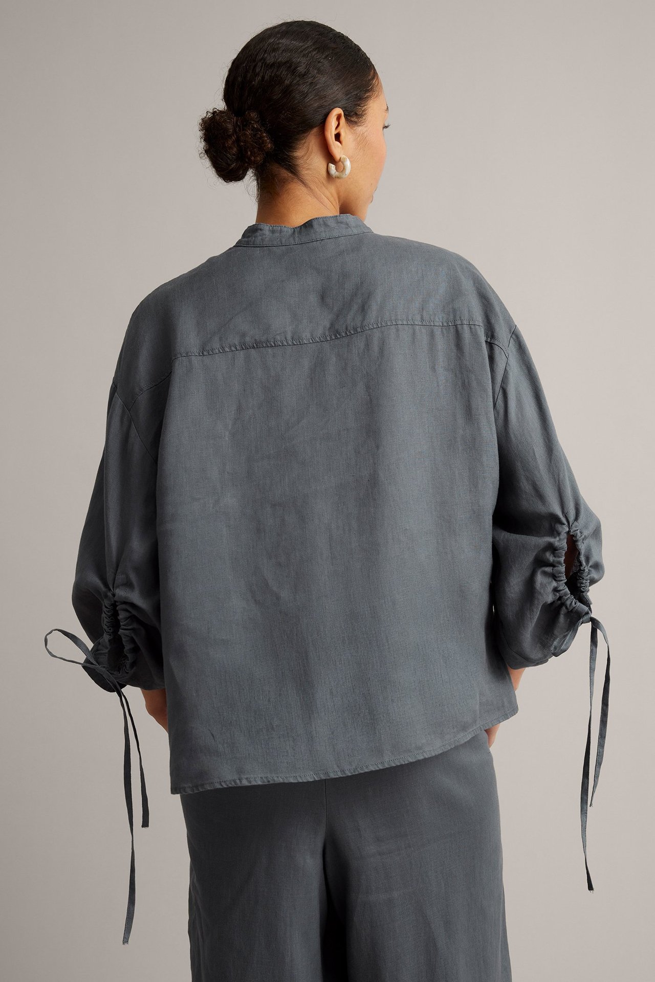 Ryggvisning av kvinne i løs mørkeblå linbluse med rynkede ermer og knytebånd, med matchende bukser.