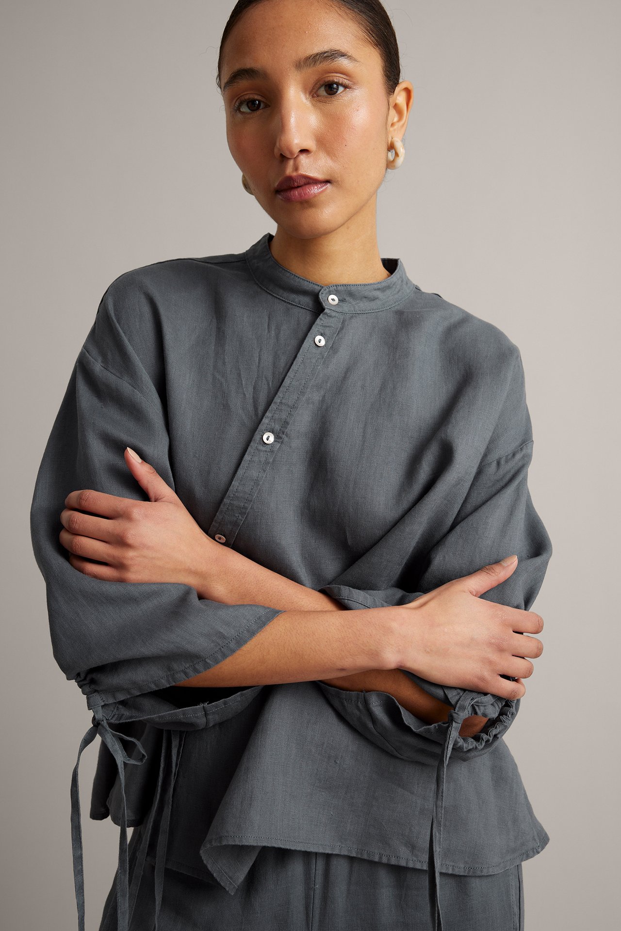 Frontutsikt: kvinne i blå linbluse med asymmetrisk knappestolpe, knytebånd i midjen og snørekanter.