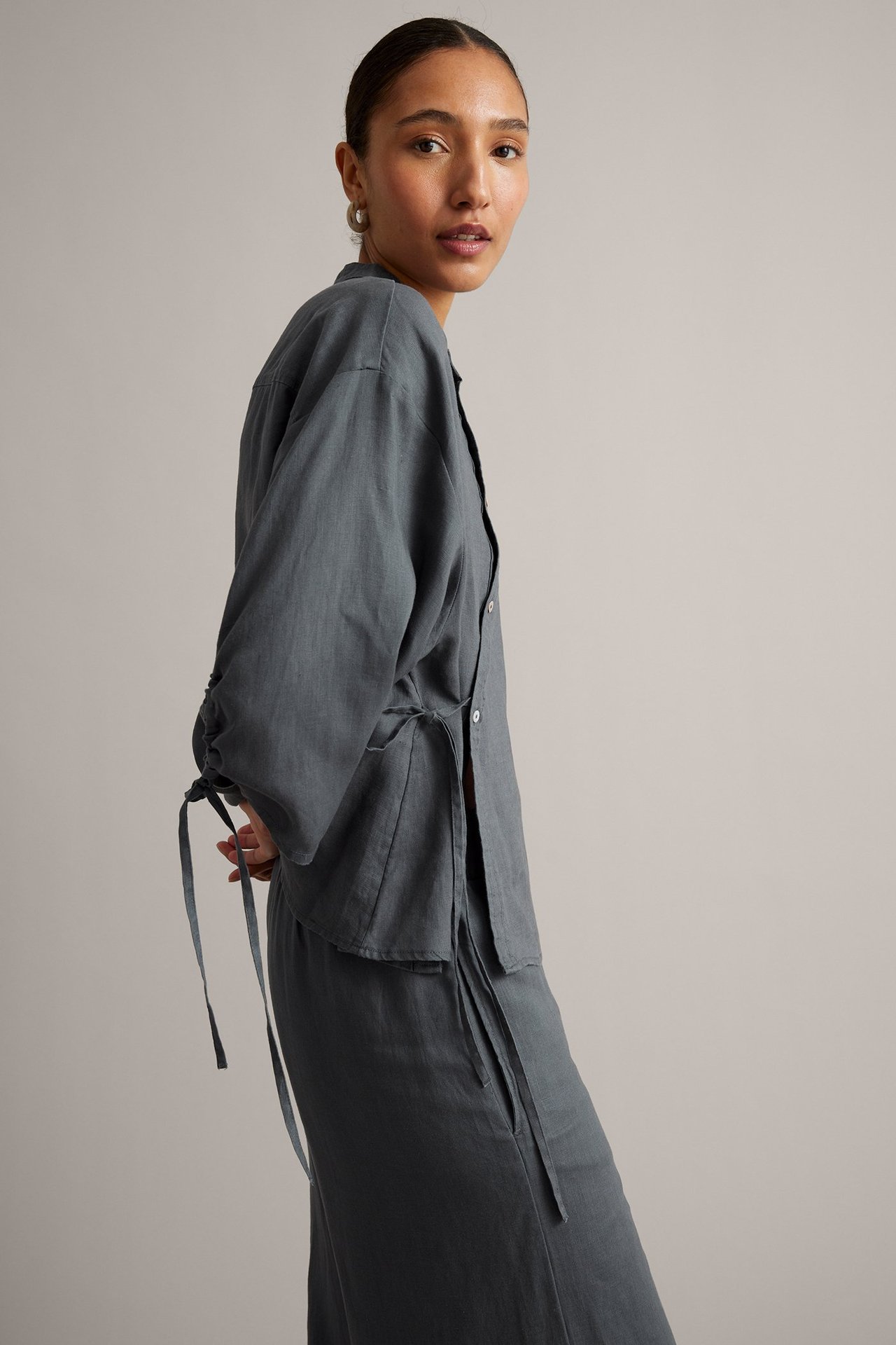 Sidebilde av kvinne i vid mørkeblå linbluse med knytebånd i midje og mansjetter, matchende skjørt og ringøredobber.