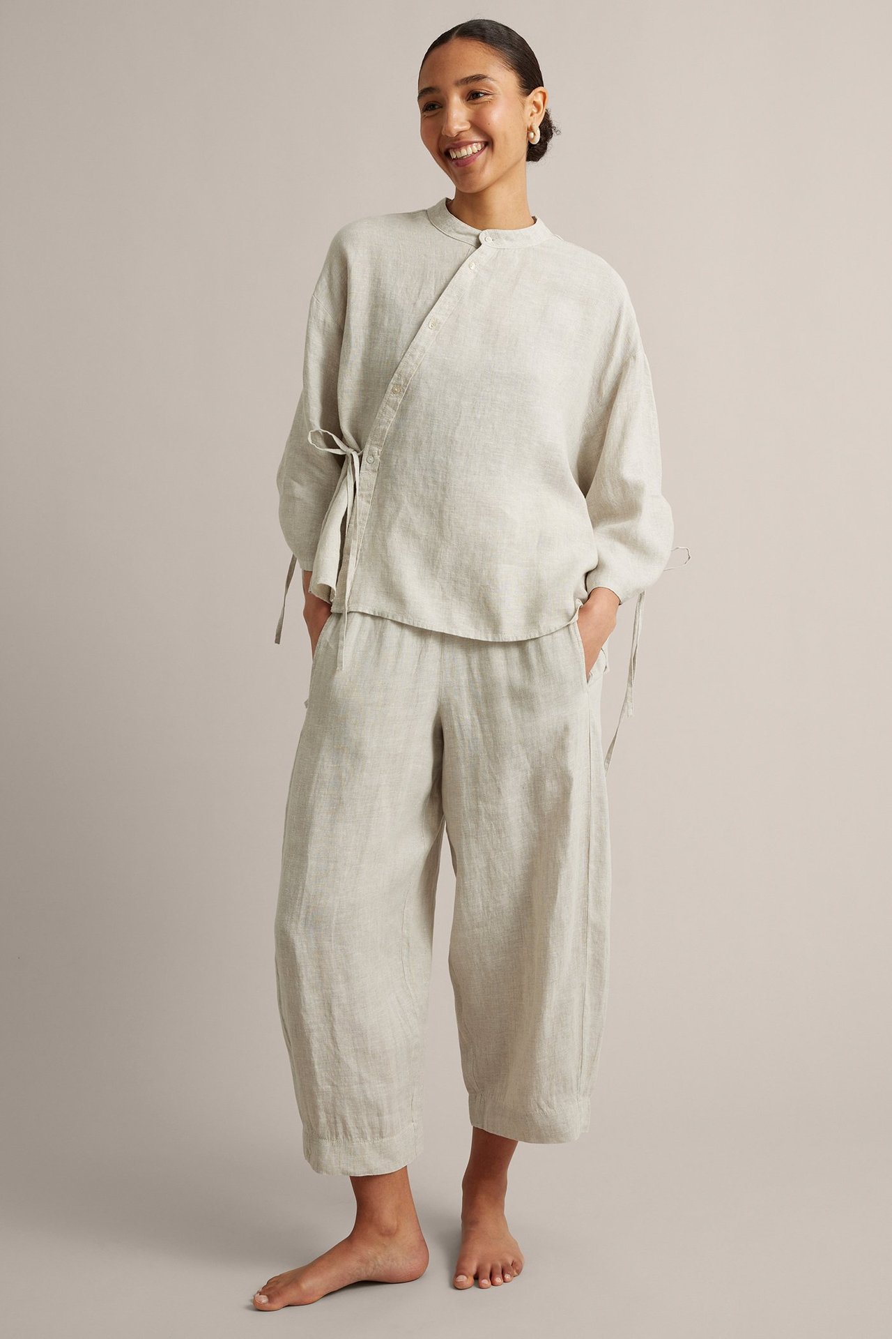 Edestäpäin nainen beigessä pellavapuserossa, epäsymmetrisellä suljennalla, ja yhteensopivissa leveälahkeisissa housuissa.