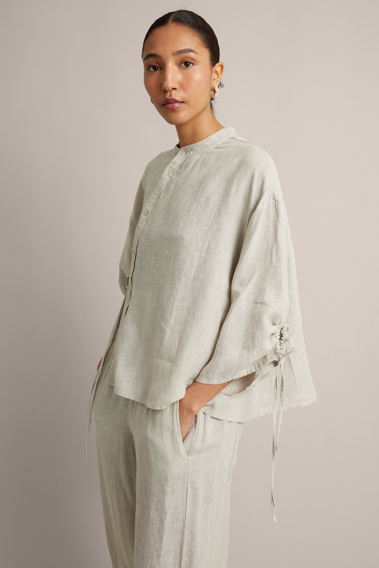 Frontbilde av kvinne i beige linbluse med asymmetrisk knepping, snøredetaljer på ermene og matchende bukser.