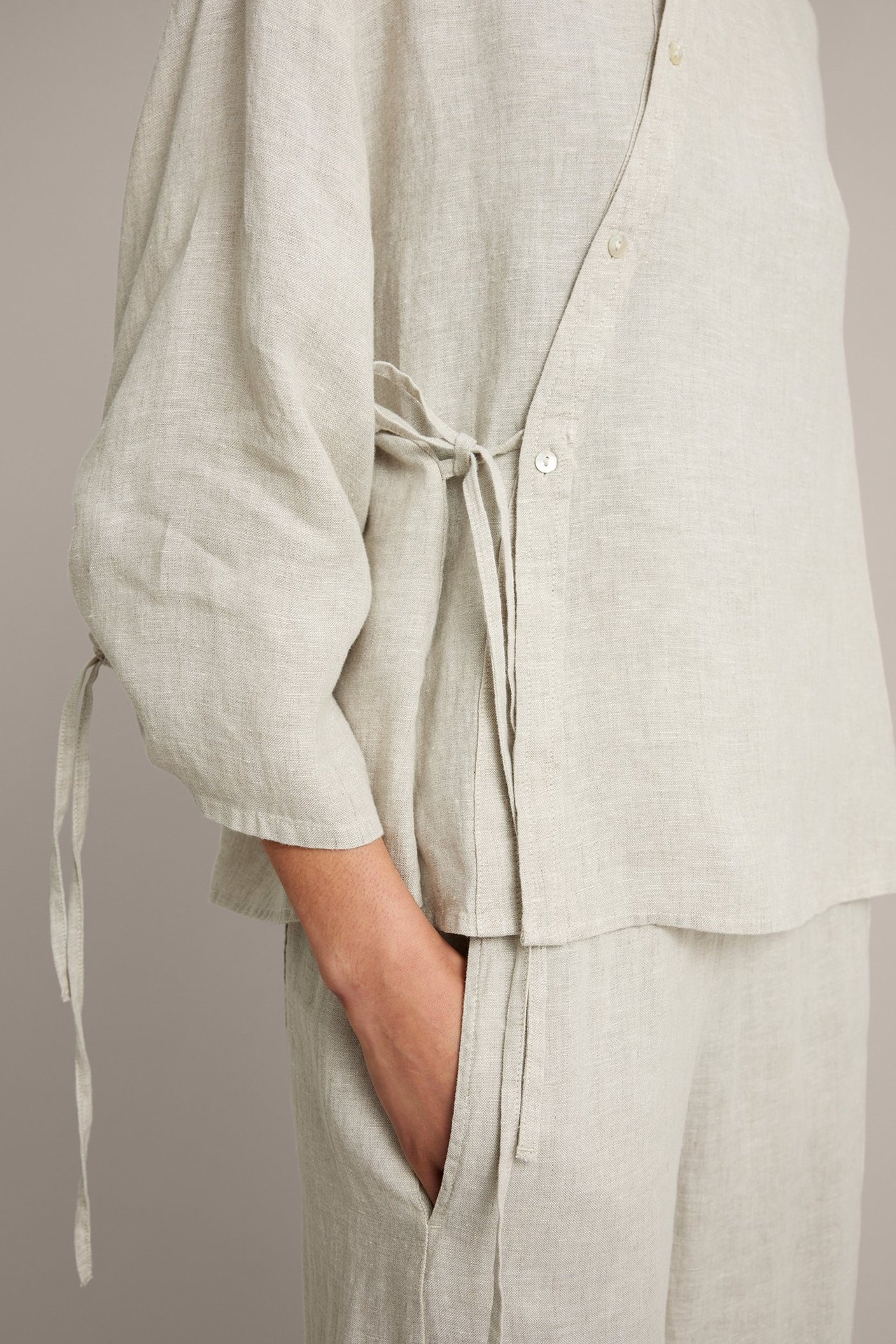 Nærbilde av kvinne i beige linbluse med skrå knapper, knytebånd i midjen og matchende bukser.