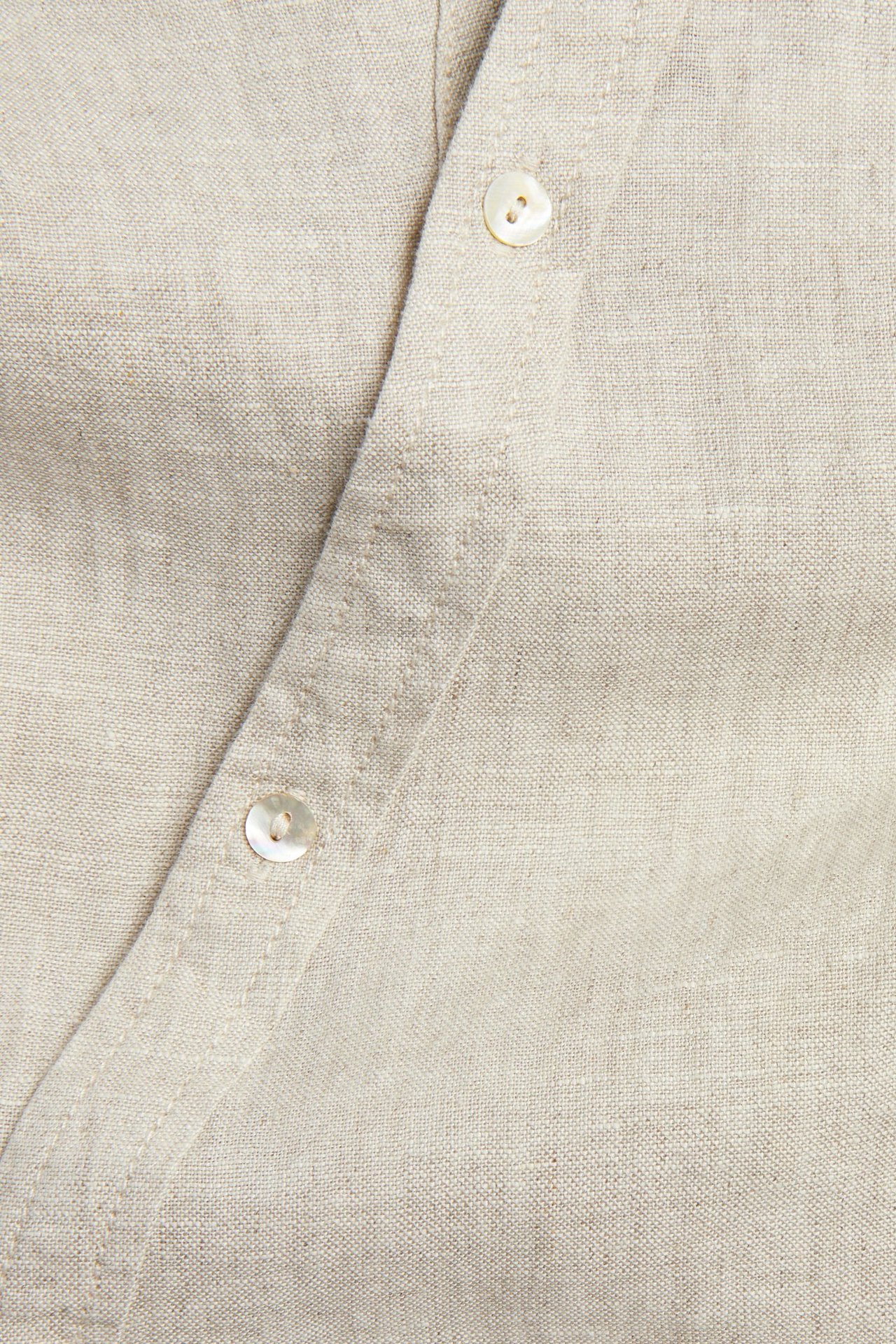 Nærbilde av en beige linbluse for dame med skrått knappestolpe og to synlige perlemorsknapper.