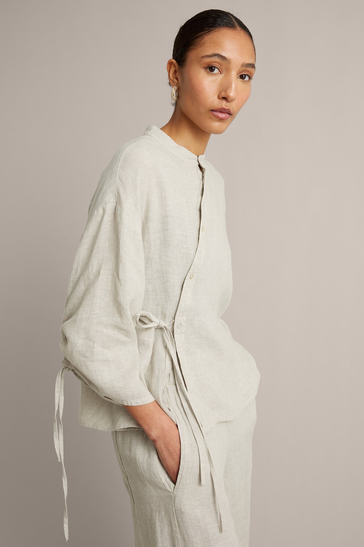 Widok z przodu kobiety w luźnej beżowej lnianej bluzce z asymetrycznym wiązaniem, do kompletu ze spodniami.