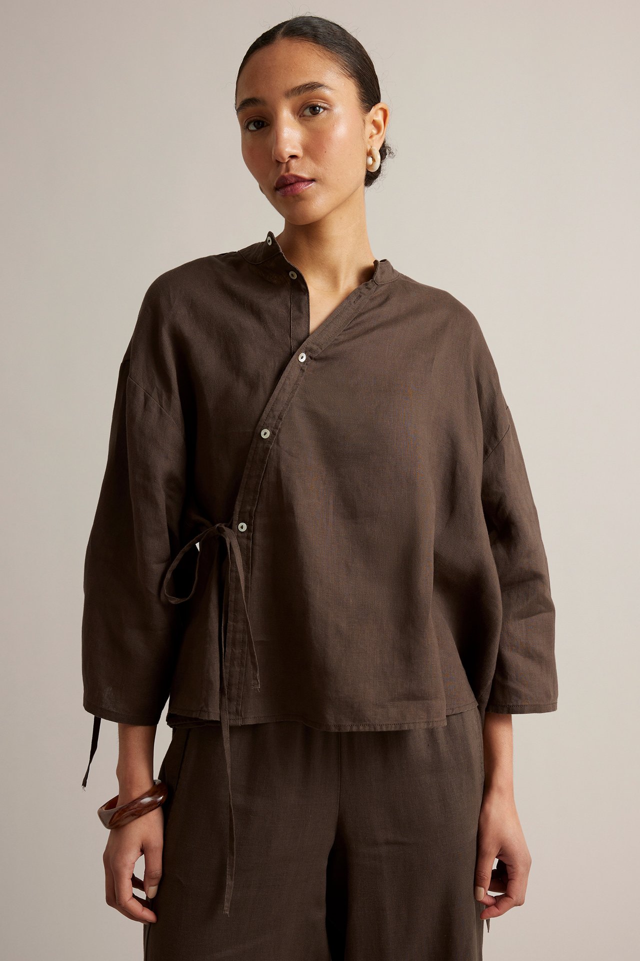 Edestä: nainen ruskeassa pellavapuserossa pystykauluksella, epäsymmetrisellä solmimisvyöllä ja yhteensopivissa housuissa.