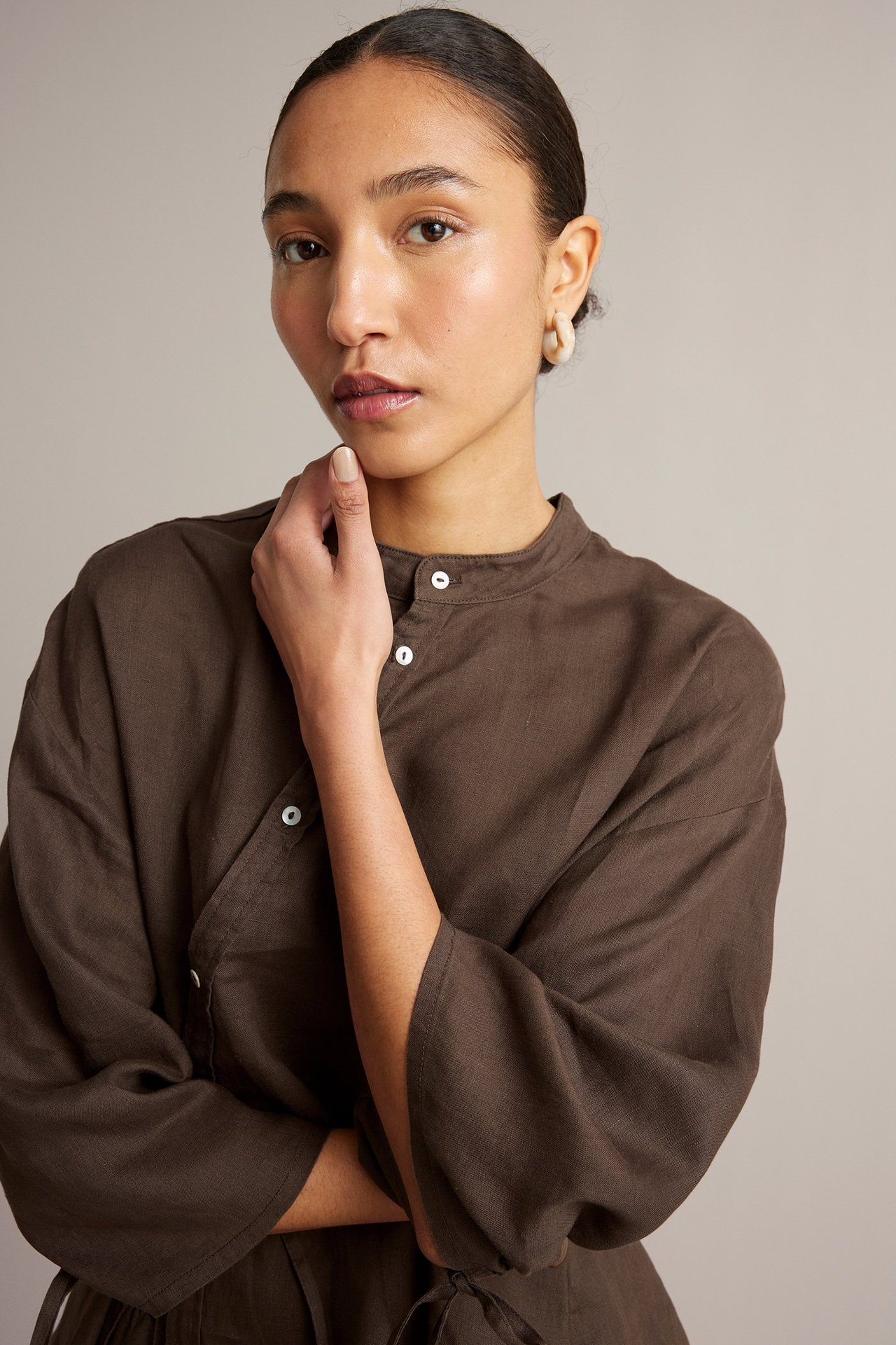 Forfra: Kvinne i mørkebrun linbluse med asymmetriske hvite knapper og lyse bøyleøredobber.