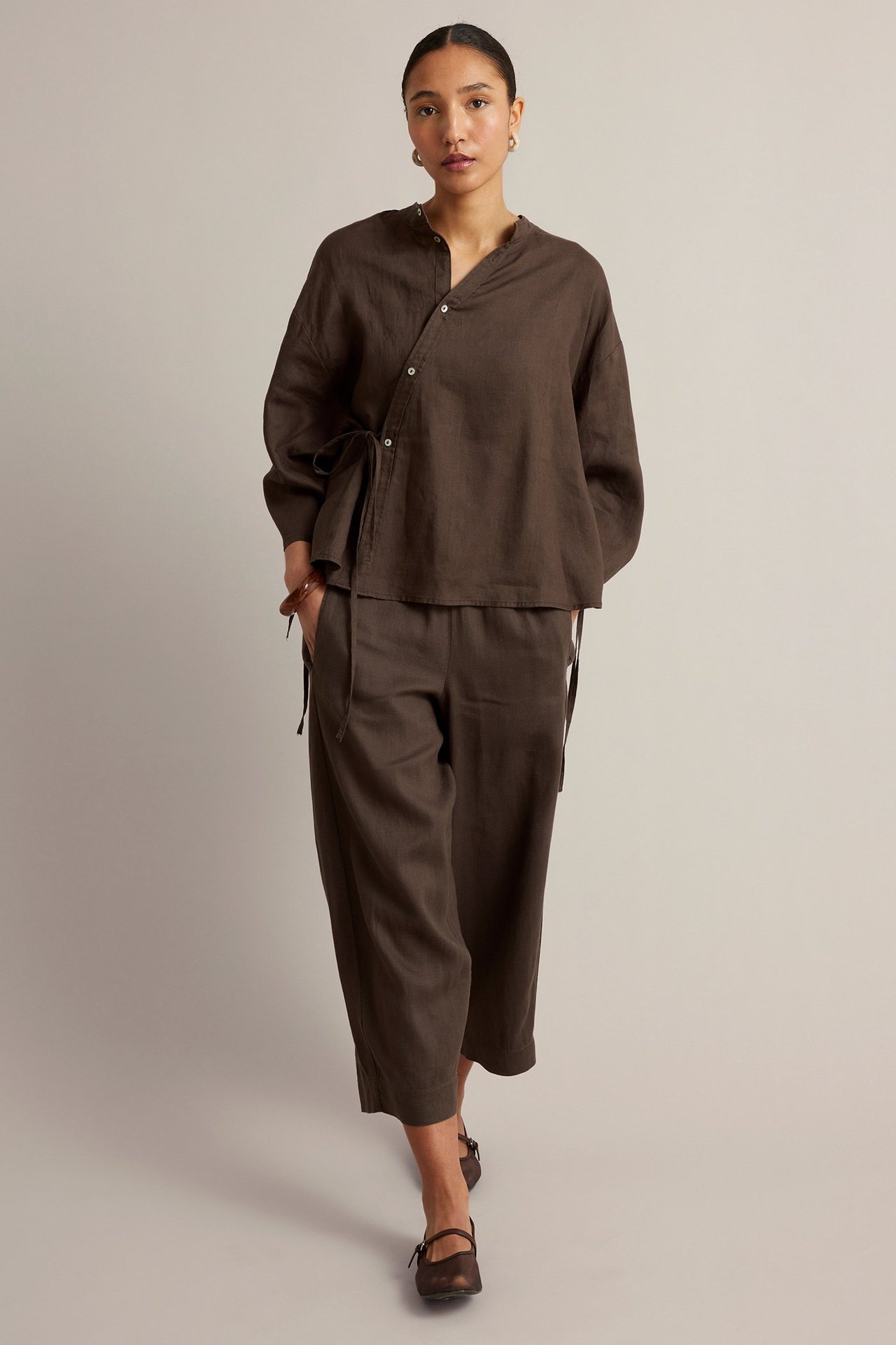 Widok z przodu: Kobieta w brązowej lnianej bluzce z wiązaniem w pasie, brązowych skróconych spodniach i butach.
