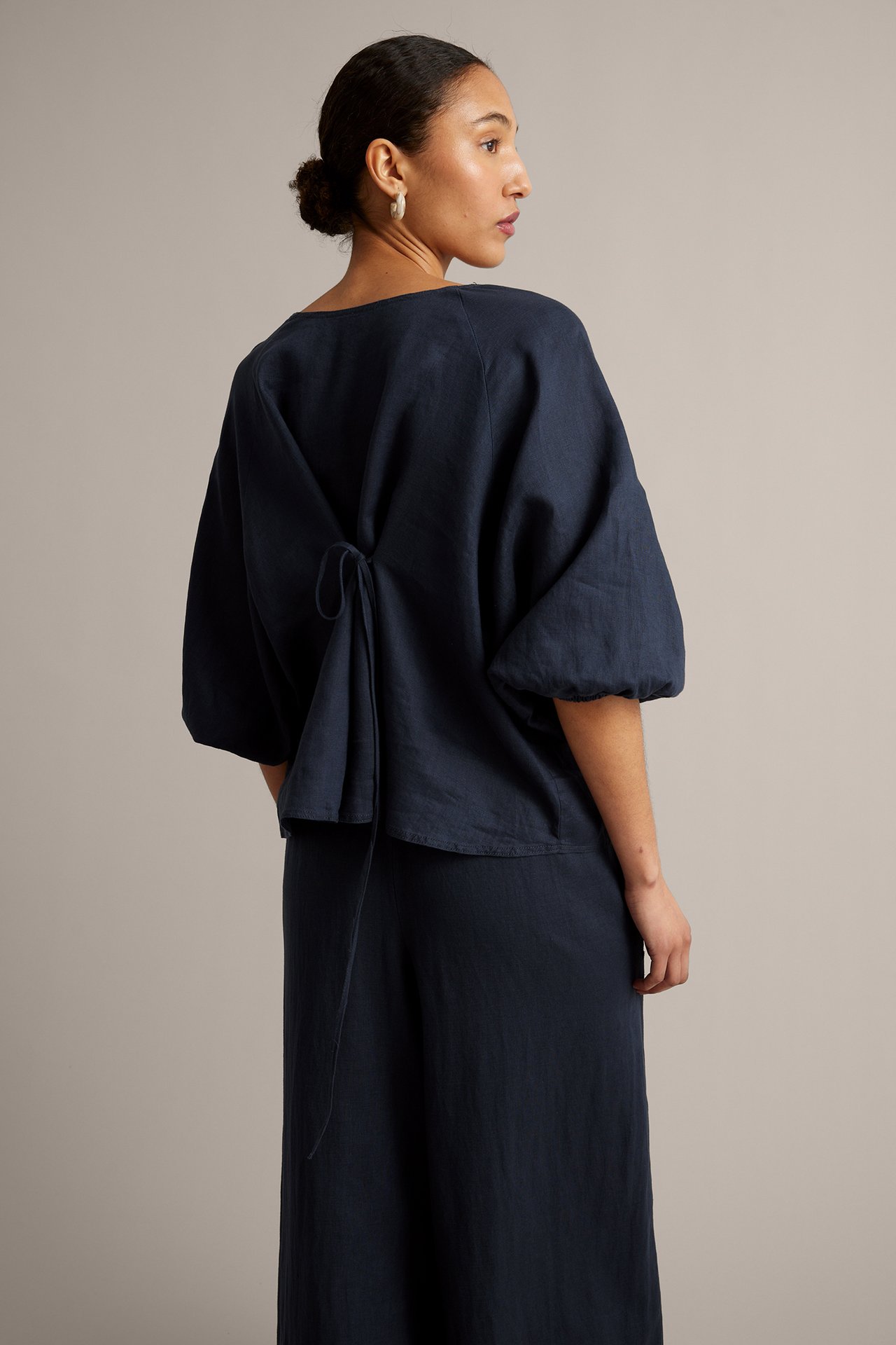 Kobieta od tyłu w ciemnoniebieskiej lnianej bluzce z wiązaniem, raglanowymi rękawami i pasującymi lnianymi spodniami.