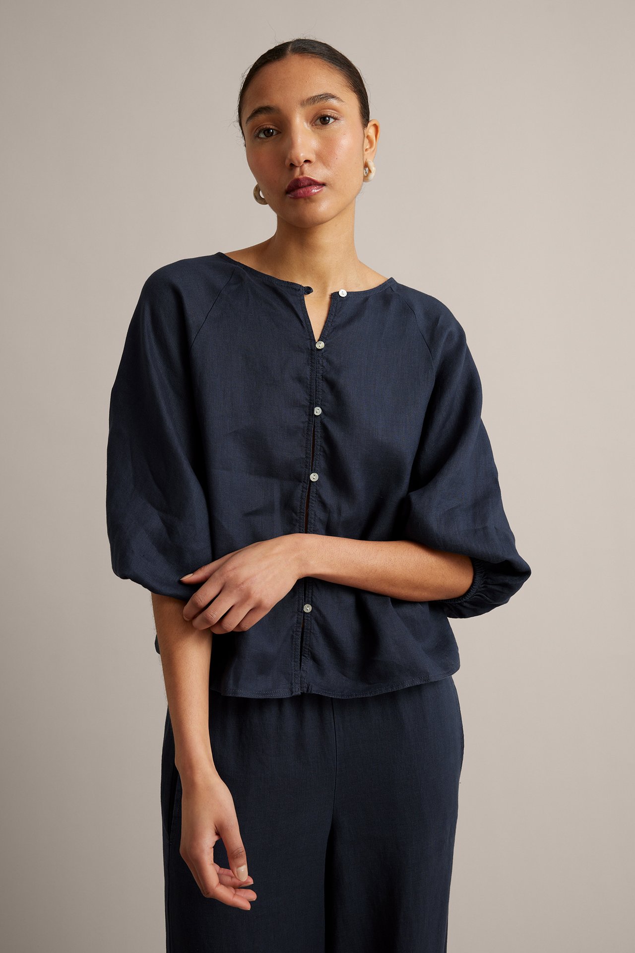 Kobieta w granatowej lnianej bluzce, białych guzikach, szerokich rękawach, pasujących spodniach, kolczykach.