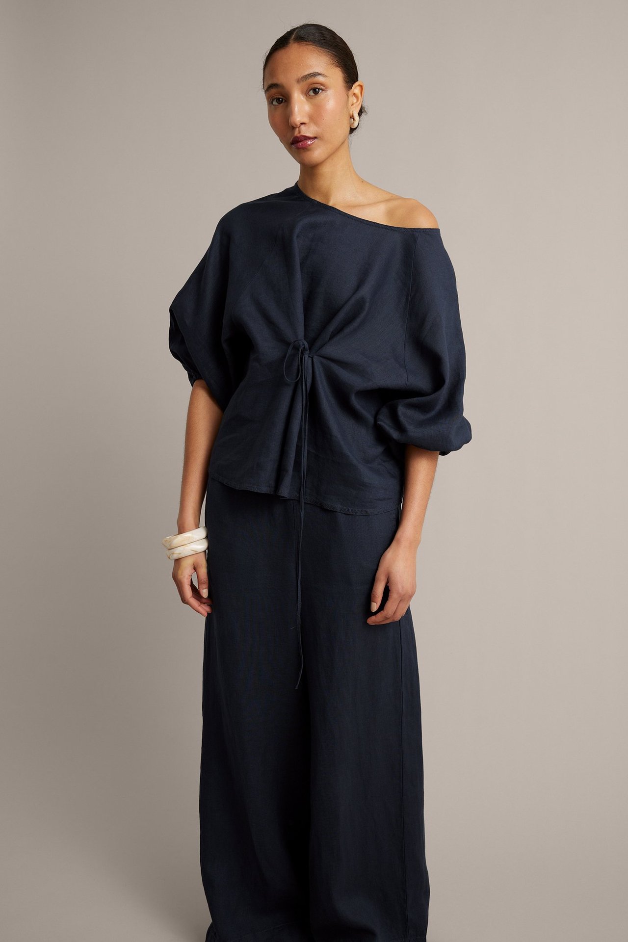 Widok z przodu: Kobieta w granatowej lnianej bluzce off-shoulder z wiązaniem, pasujących spodniach i kremowych bransoletkach.