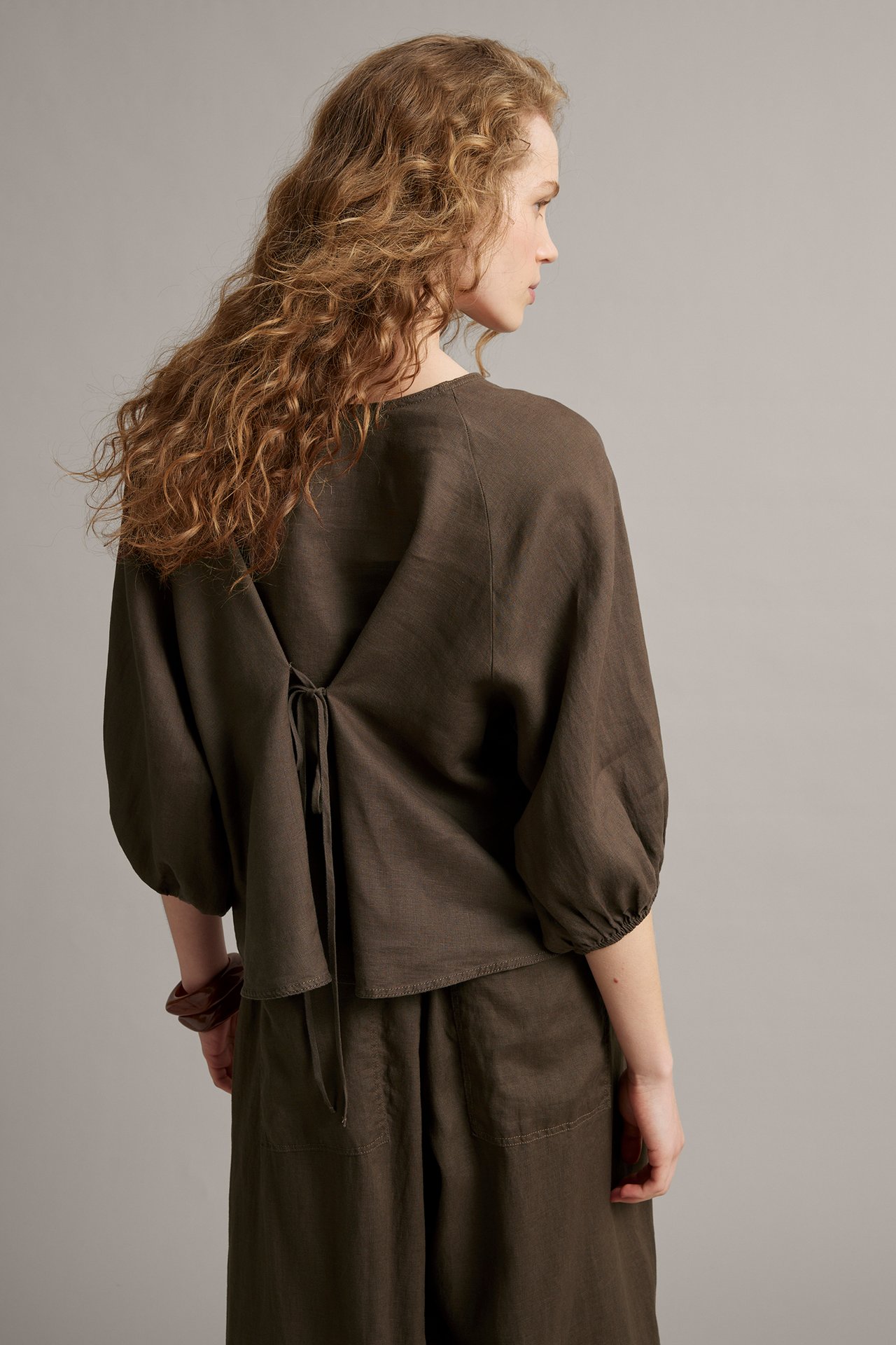 Widok z tyłu kobiety w brązowej lnianej bluzce z wiązaniem z tyłu i pasujących spodniach, noszącej brązową bransoletkę.