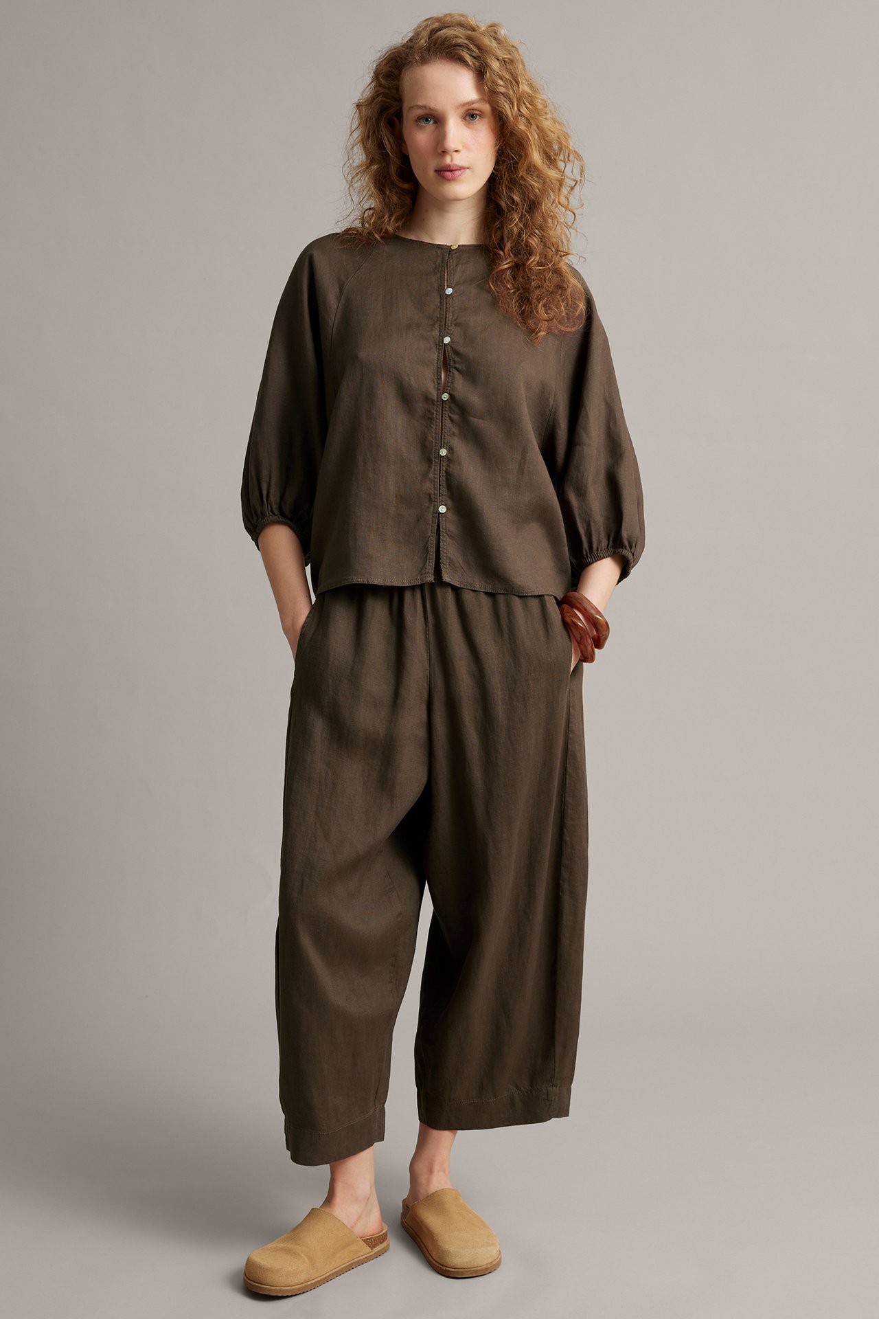 Framsida: Kvinna i brun linneblus med vita knappar, matchande vida byxor, beiga tofflor och armband.