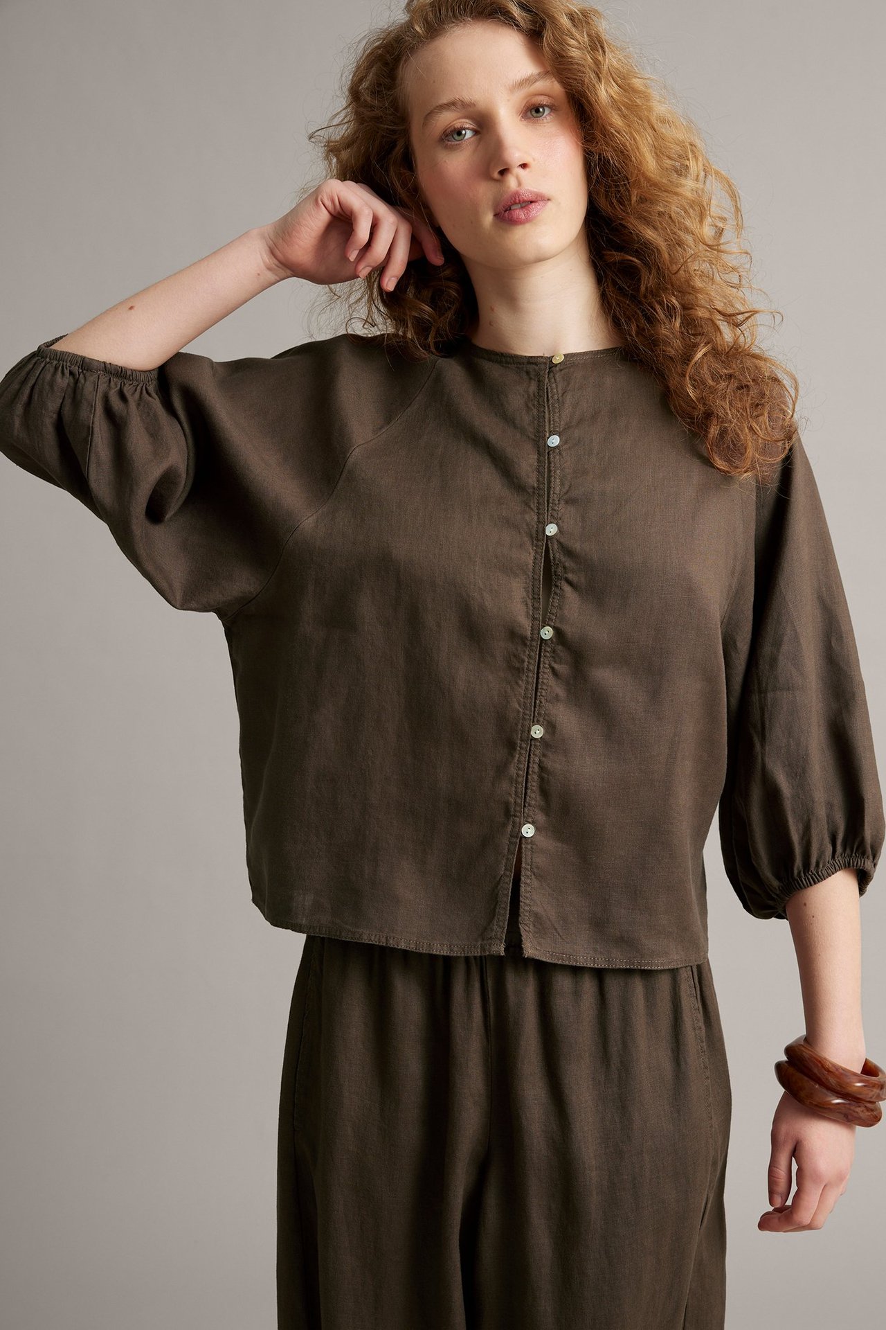 Z przodu: Kobieta w brązowej lnianej bluzce z guzikami, dopasowanych spodniach i brązowych bransoletach.