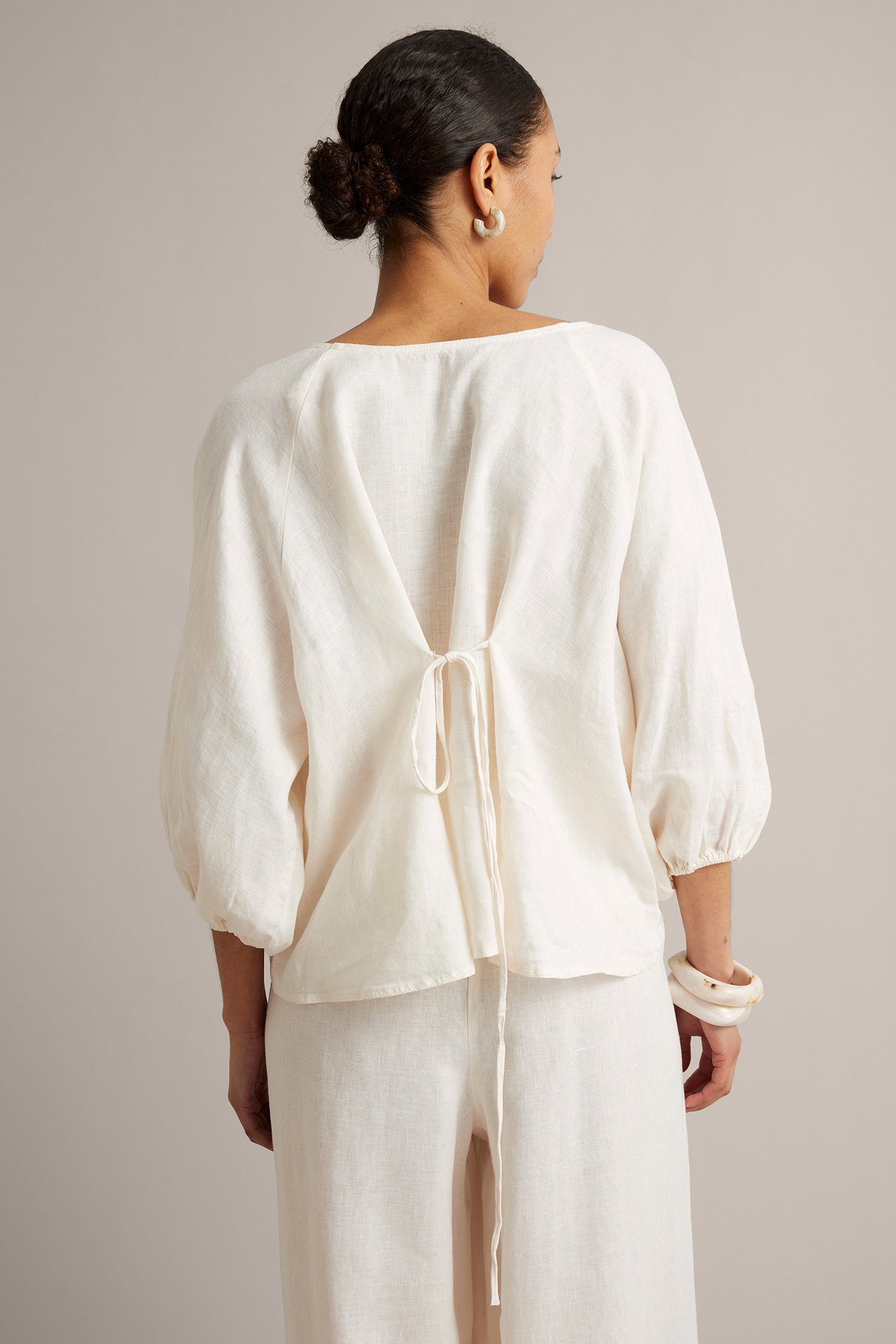 Widok z tyłu: kobieta w luźnej białej bluzce lnianej z wiązaniem, pasujących spodniach, kremowych kolczykach i bransoletce.