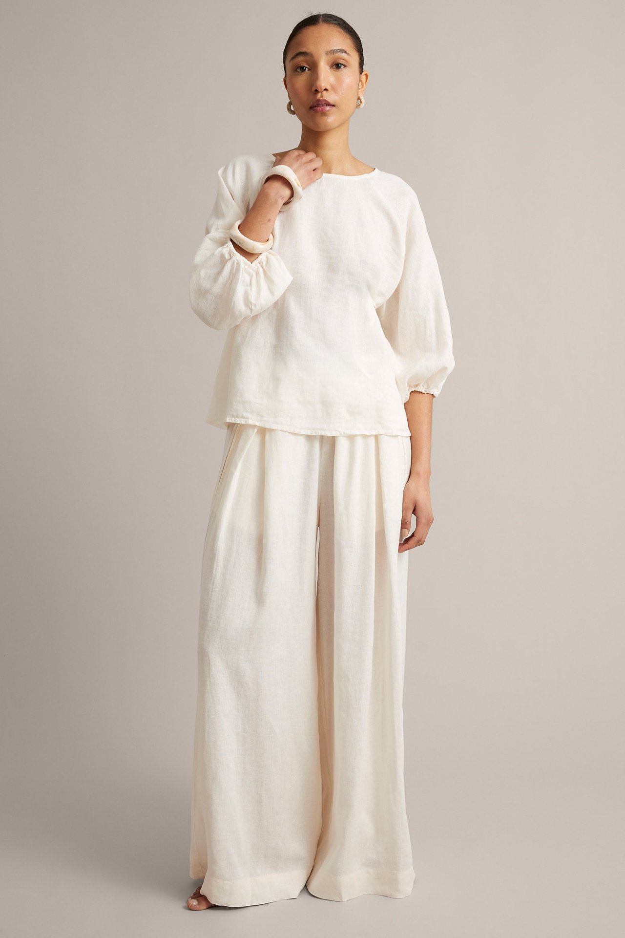 Nainen valkoisessa pyöreäkauluksisessa pellavapuserossa, jossa volyymihihat, yhteensopivissa leveissä housuissa ja valkoisessa rannekorussa.