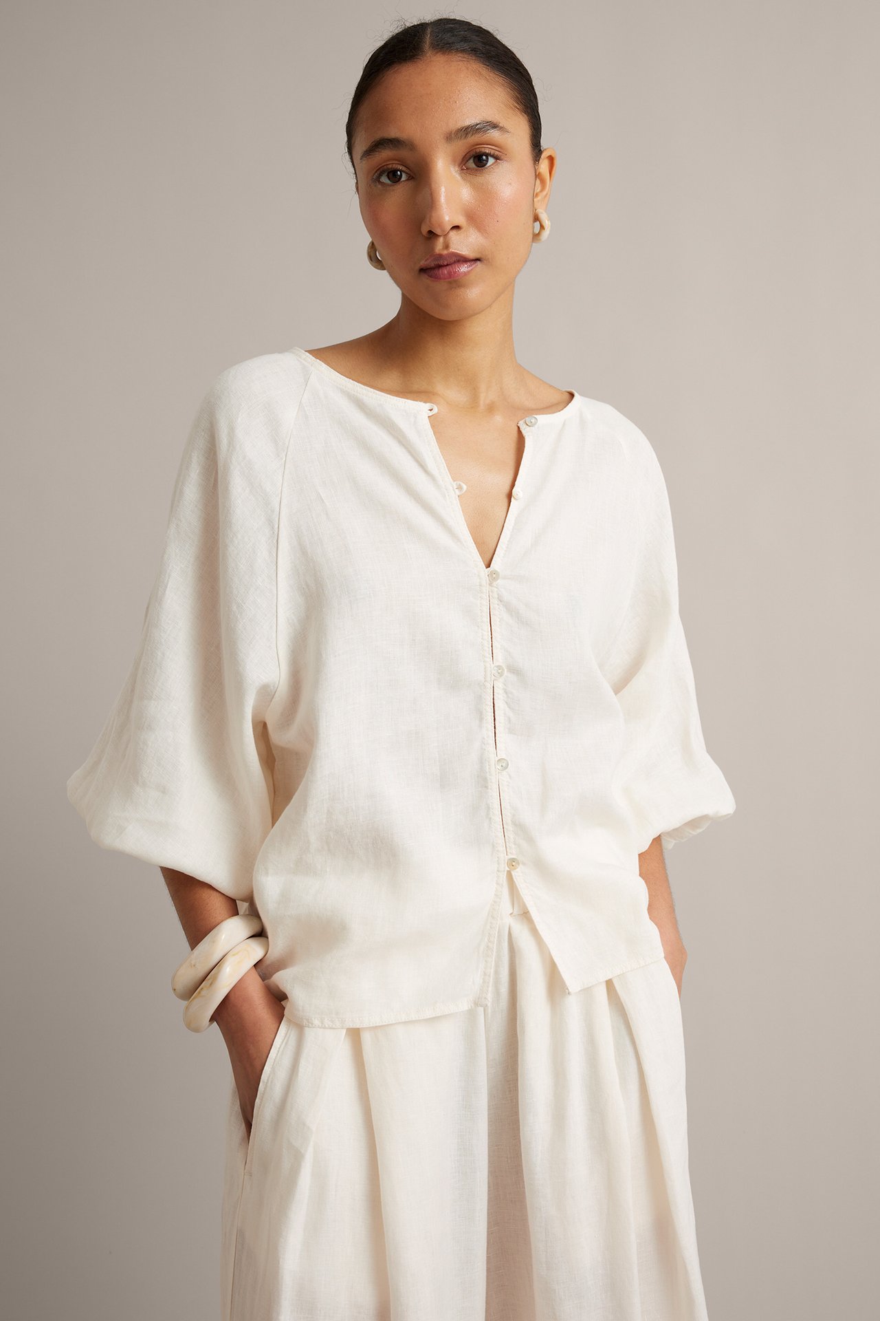 Widok z przodu: Kobieta w białej lnianej bluzce z szerokimi rękawami raglan, pasujące spodnie, bransoletki i kolczyki.
